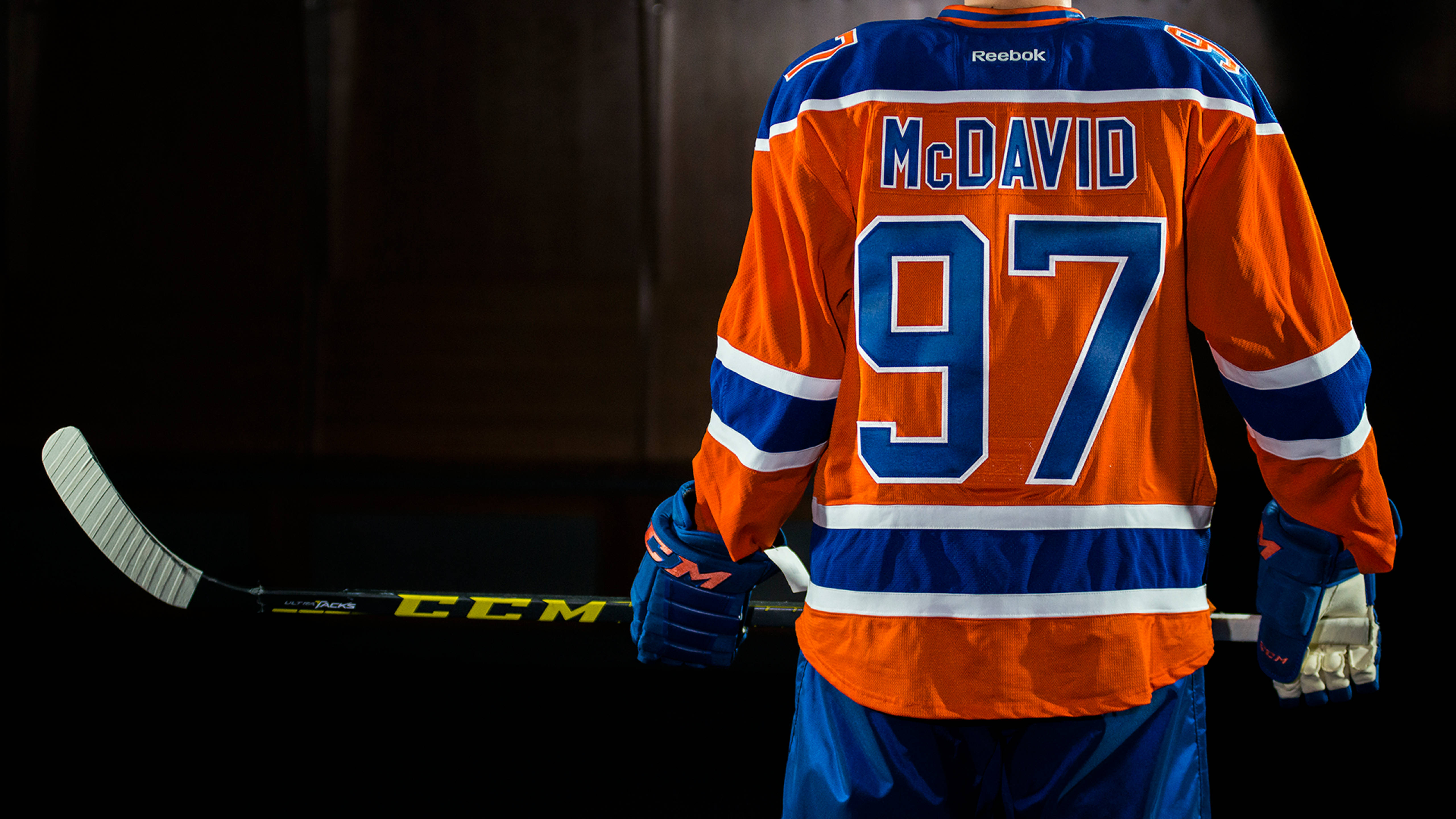 Oilers 桌布 壁紙 — 一名身穿橙藍色球衣、號碼97的曲棍球運動員站在深色背景前，手持球棍。