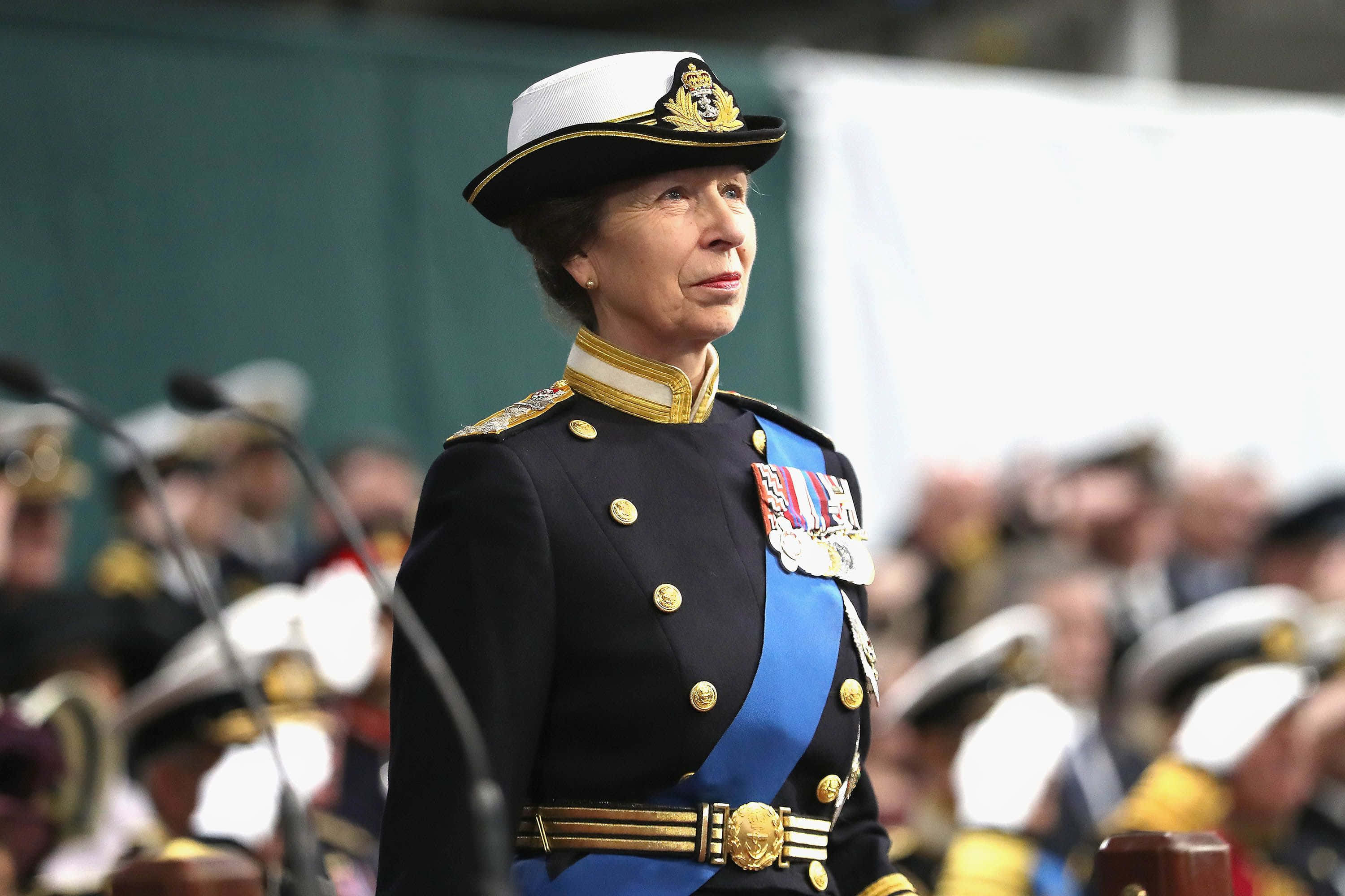 軍事動圖 桌布 壁紙 — 一名身穿正式海軍制服、佩戴勳章和金色飾邊的女性，在麥克風和模糊的與會者背景下，莊嚴地站在典禮活動中。