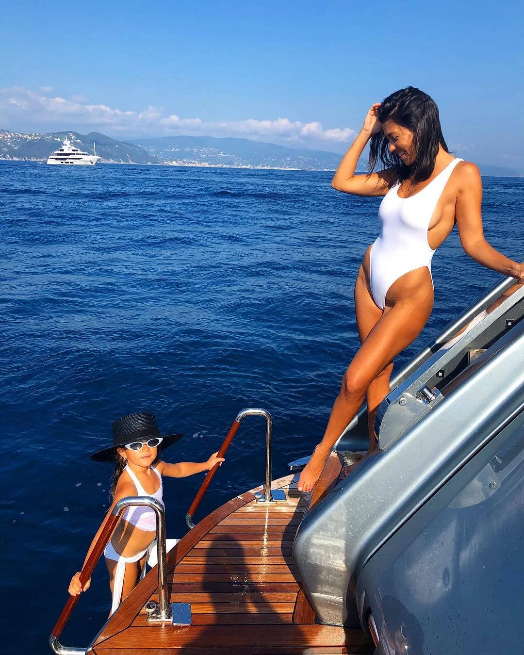 Kourtney 桌布 壁紙 — 一名身穿泳衣的女子和小孩在清澈的天空下，以深藍色的大海和遠山為背景，踏上遊艇甲板。