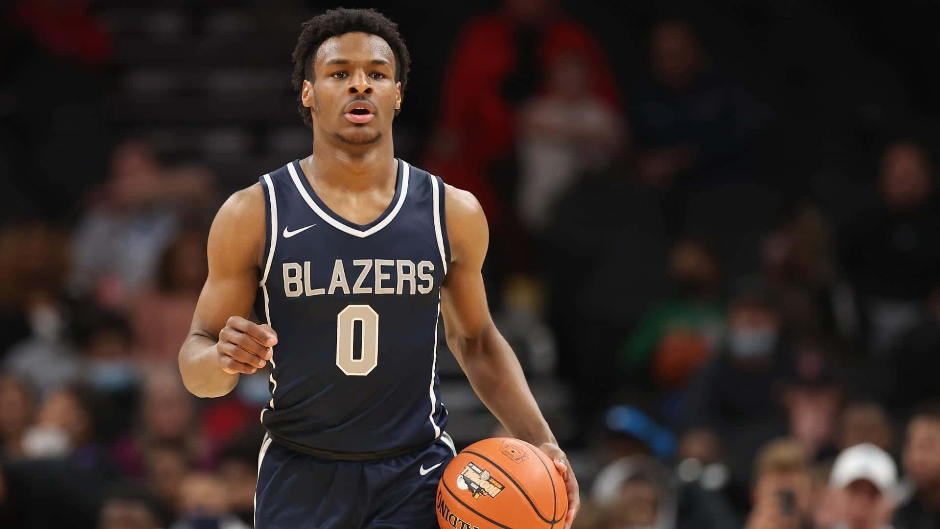 Bronny James 桌布 壁紙 — 一名身穿海軍藍 Blazers 球衣的籃球運動員在比賽中運著橙色籃球，背景是模糊的觀眾。