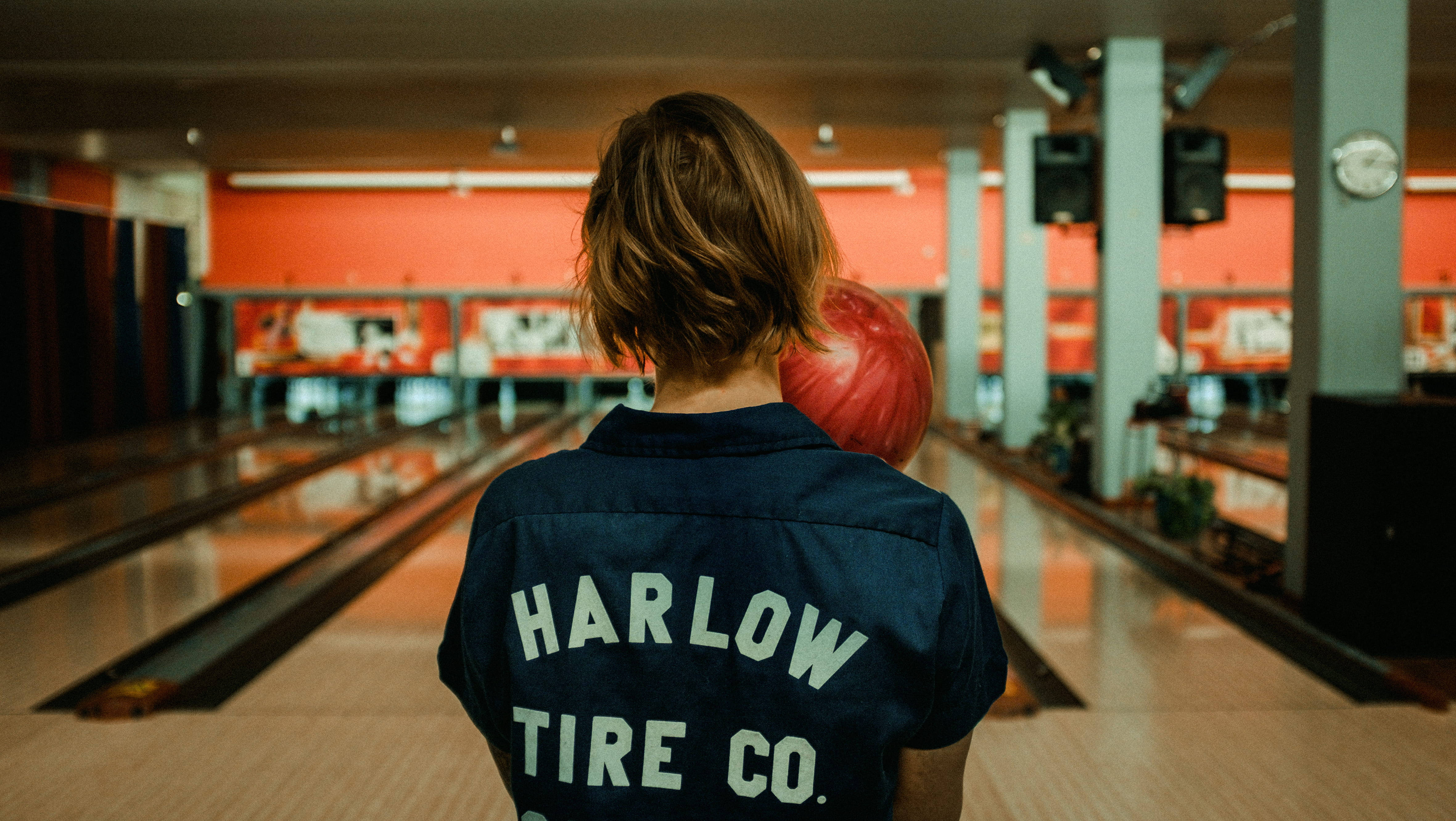 保齡球 桌布 壁紙 — 一名身穿海軍藍 'Harlow Tire Co.' 襯衫的人，在燈光溫暖的保齡球館球道起點拿著一個紅色保齡球。