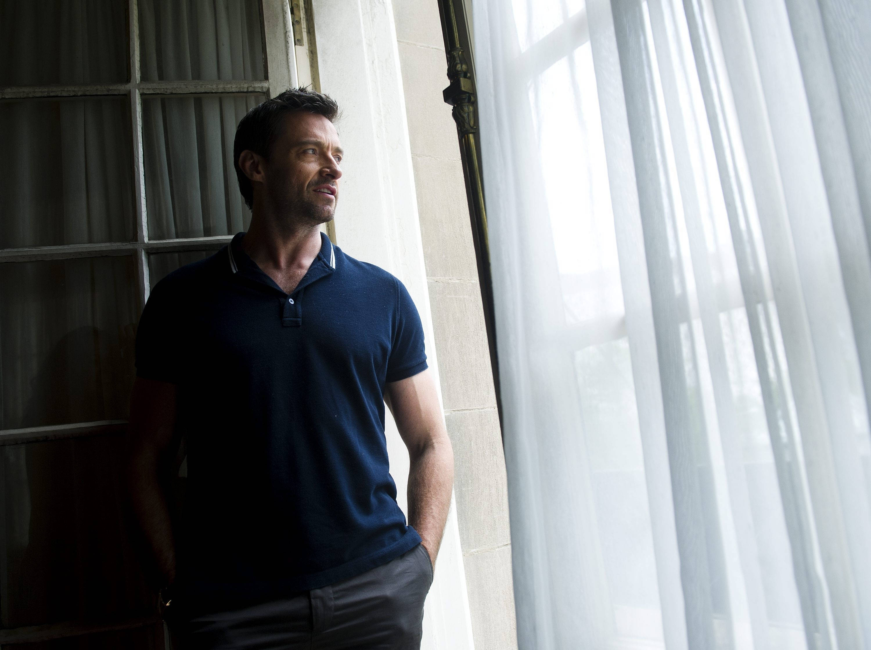 Hugh Jackman 桌布 壁紙 — 一名身穿海軍藍Polo衫的男子站在窗邊，窗簾輕薄，在柔和自然光中若有所思地向外凝望。