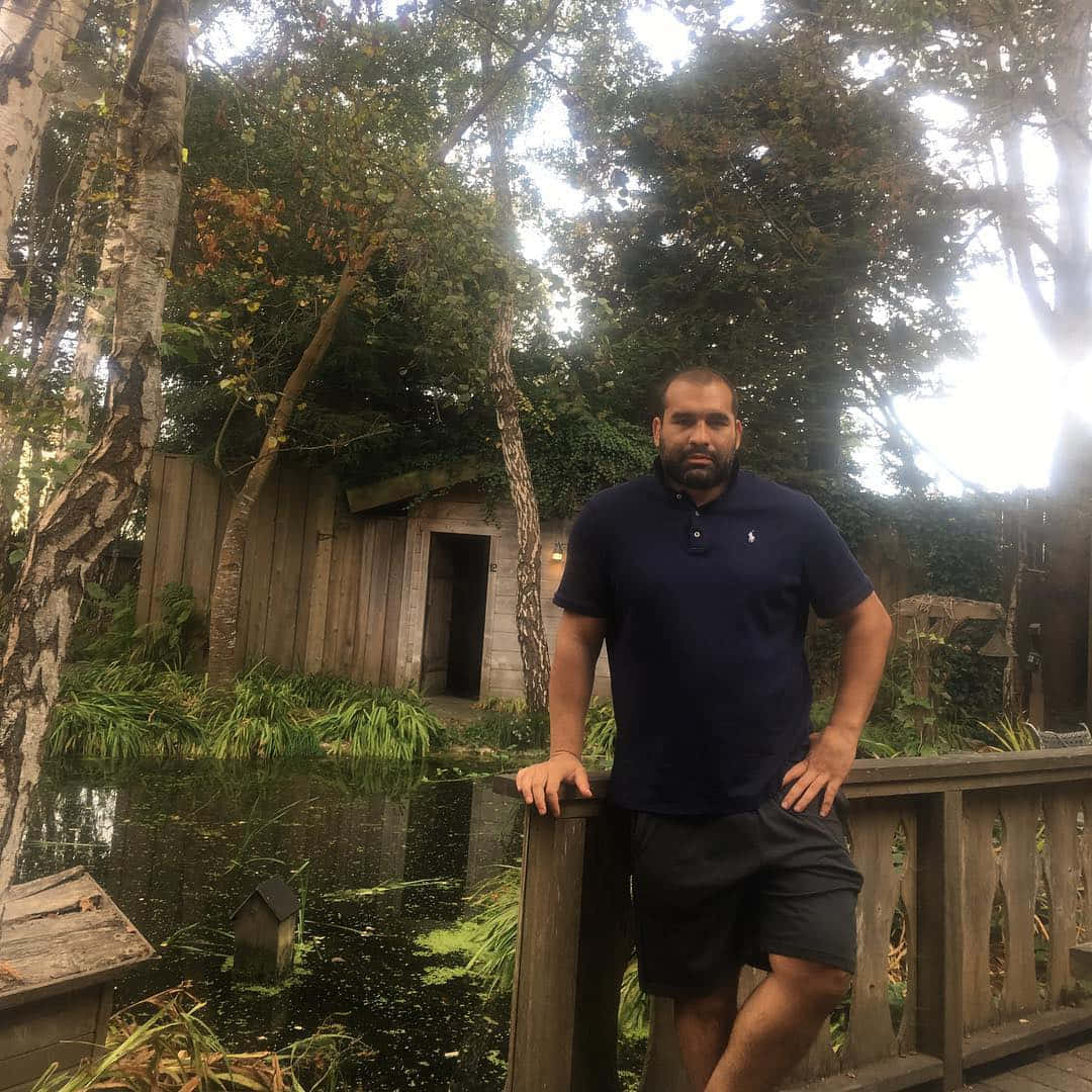 Celebrity 桌布 壁紙 — 一名身穿海軍藍Polo衫的男子靠在木製甲板欄杆上，旁邊是一個被茂密綠植和樹木環繞的寧靜池塘。