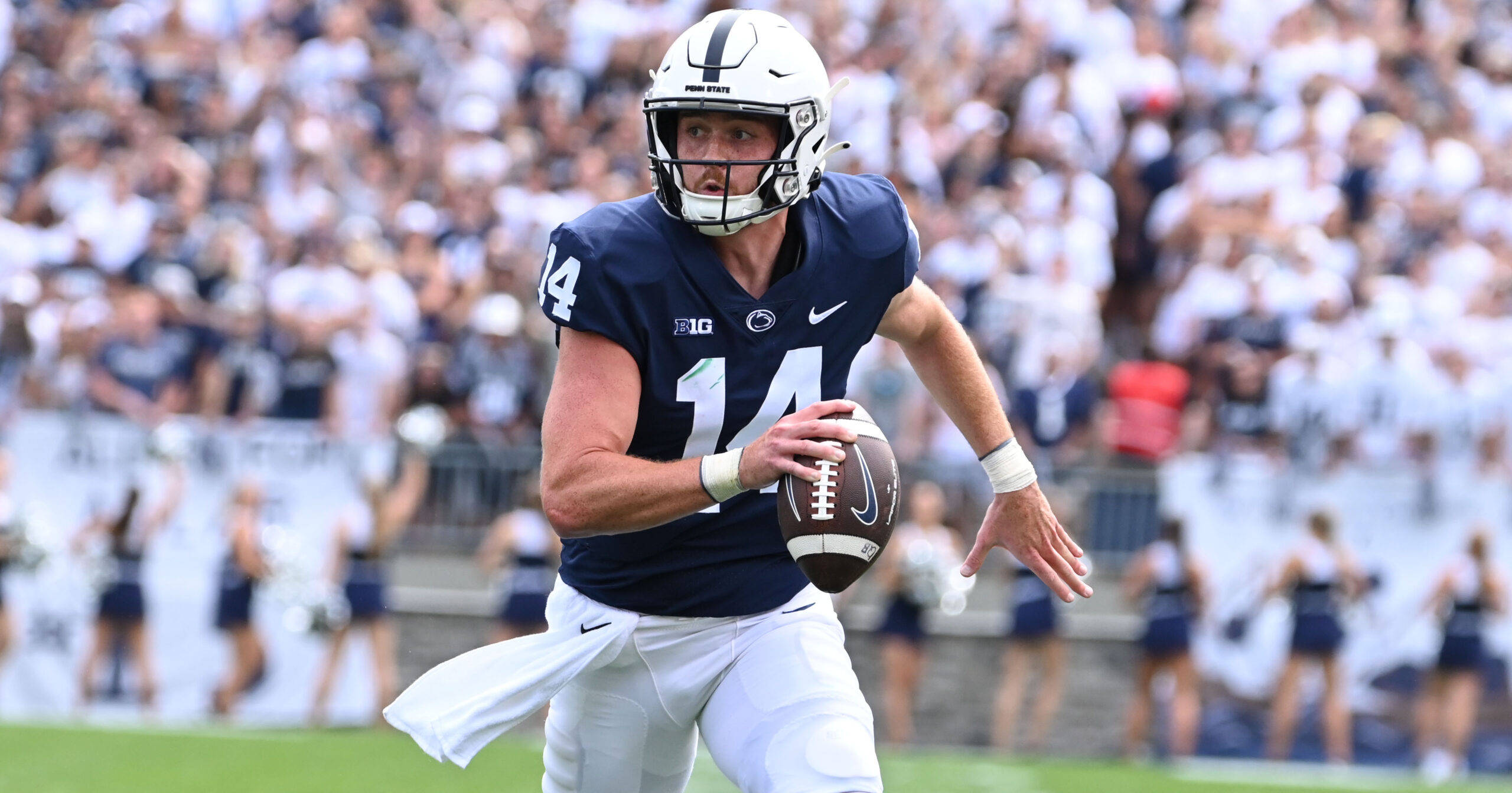 Penn State 桌布 壁紙 — 一名身穿海軍藍和白色球衣的美式足球四分衛在白天比賽中準備傳球，背景是模糊的人群。