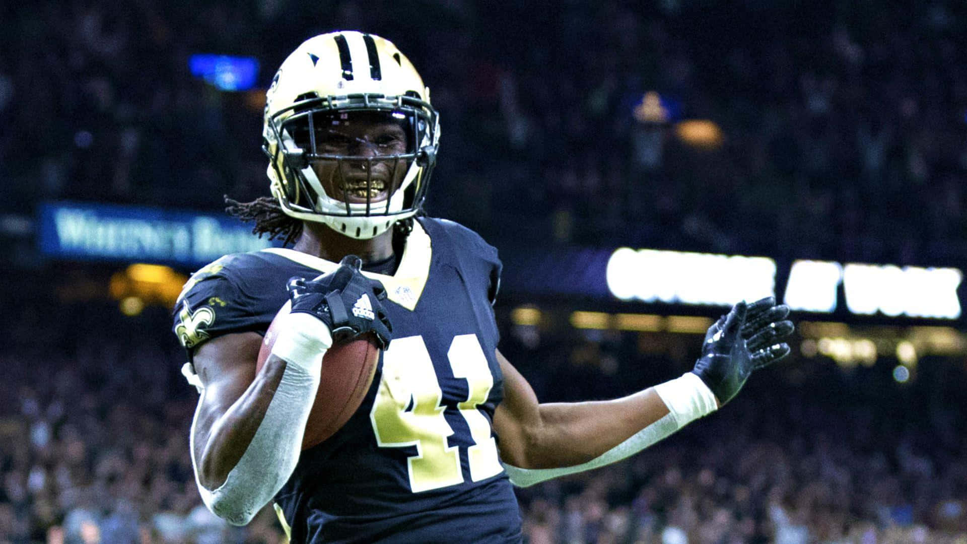 Alvin Kamara 桌布 壁紙 — 一名身穿海軍藍和金色球衣的美式足球運動員在體育場燈光下拿著足球慶祝。