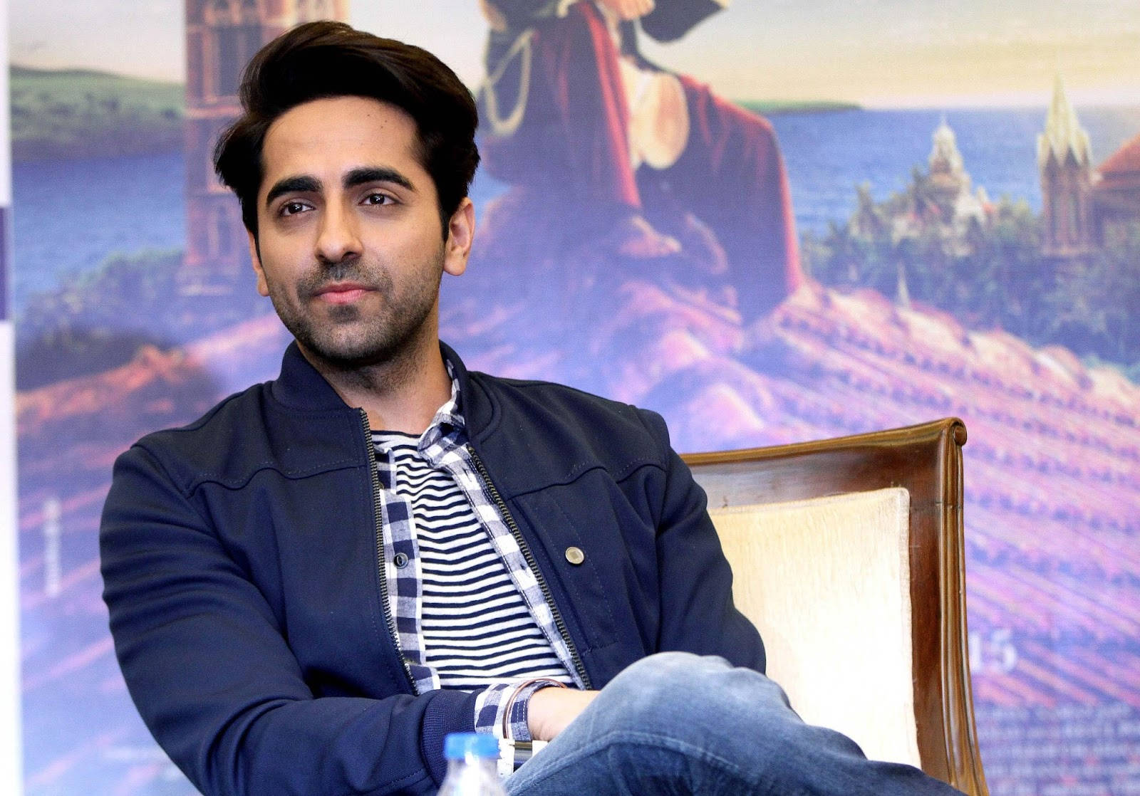 ayushmann khurrana 桌布 壁紙 — 一名身穿海軍藍外套和條紋襯衫的男子，在色彩繽紛的電影海報背景前，平靜地坐在木椅上。