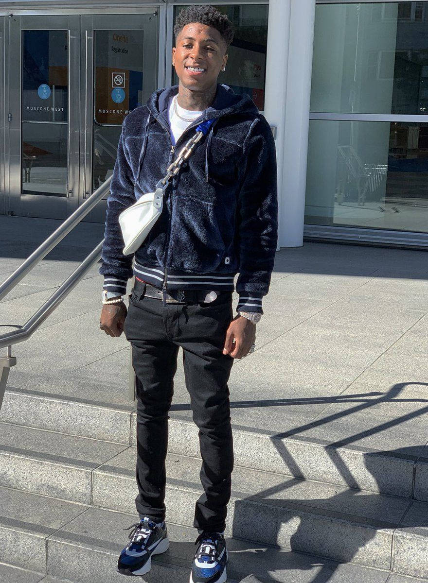 Youngboy 桌布 壁紙 — 一名身穿海軍藍抓絨連帽衫和黑色褲子的年輕男子，在玻璃建築入口附近的戶外石階上微笑站立。