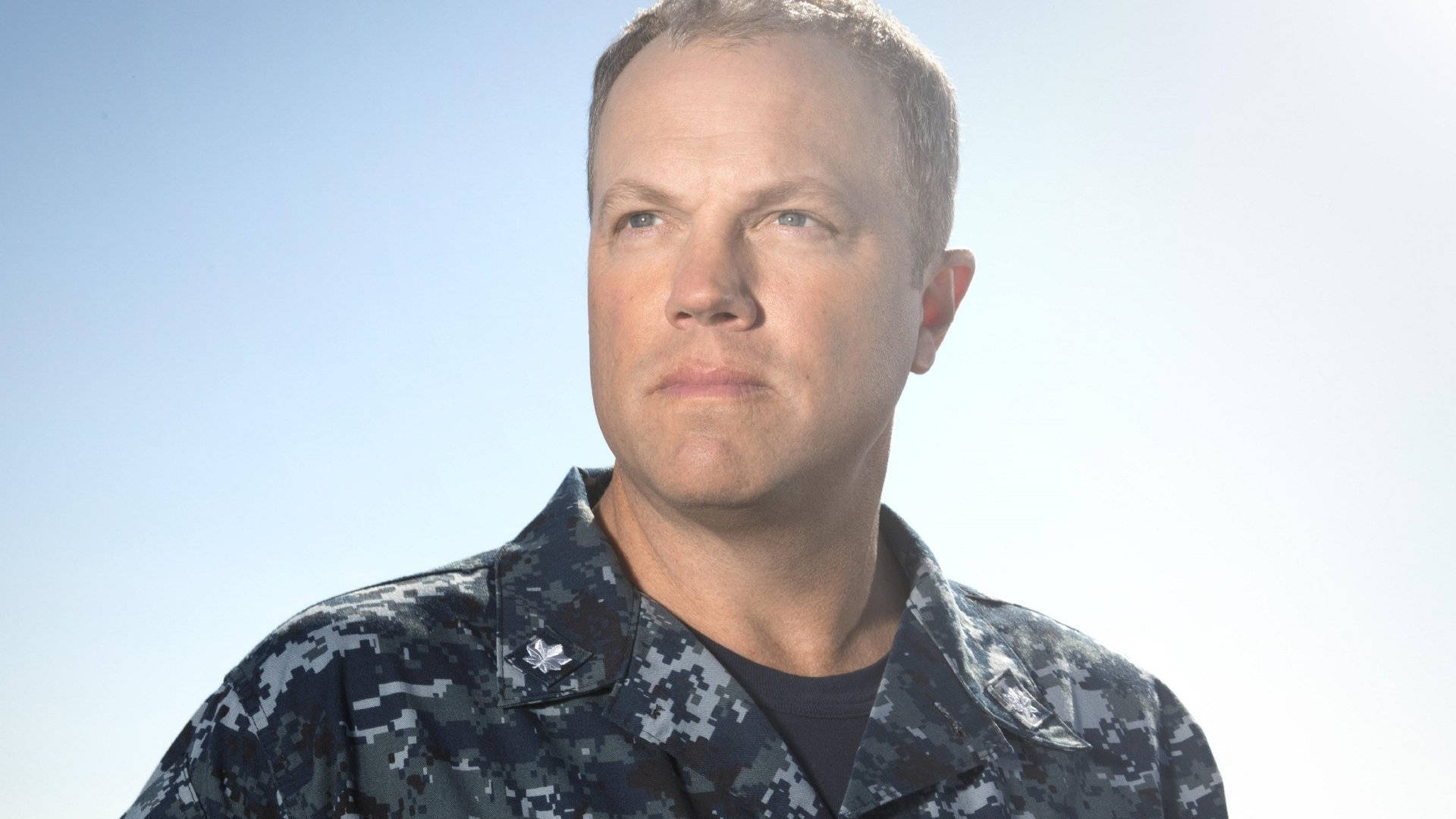 adam baldwin 桌布 壁紙 — 一名身穿海軍藍數位迷彩制服的男人，神情嚴肅地凝視著清澈的藍天。