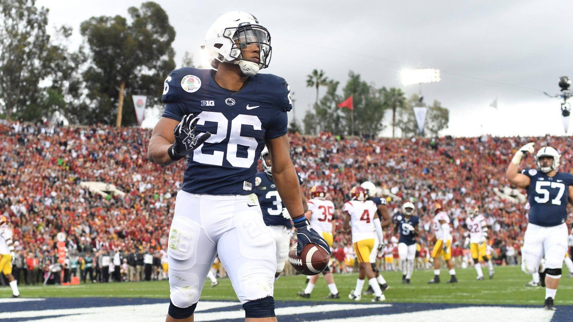 Saquon Barkley 桌布 壁紙 — 一名身穿海軍藍球衣、號碼26的足球員在陰天、擁擠的球場上拿著球向上看。
