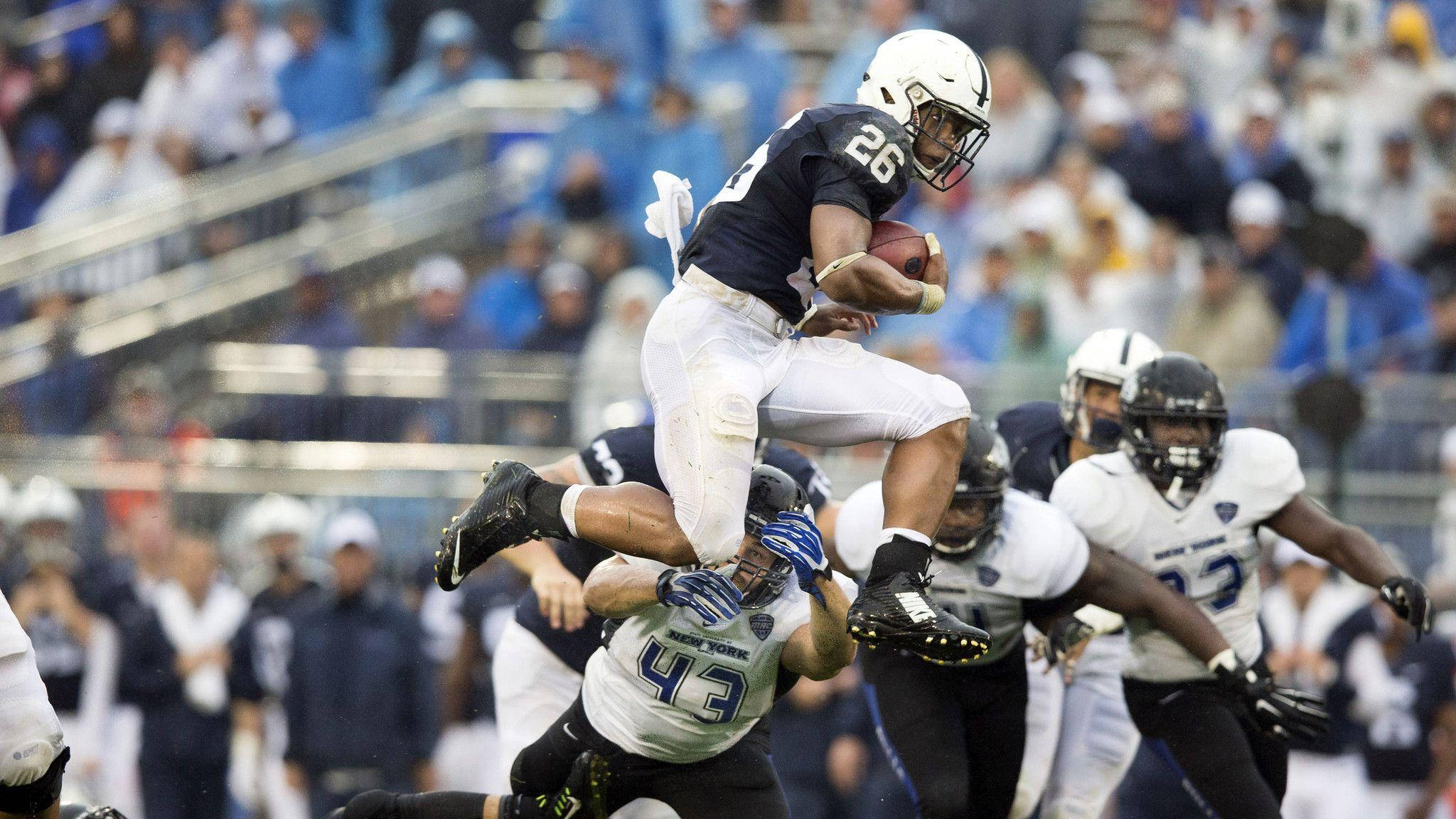 Saquon Barkley 桌布 壁紙 — 一名身穿海軍藍球衣的足球員在日間比賽中抱著球跳過一名擒抱者。