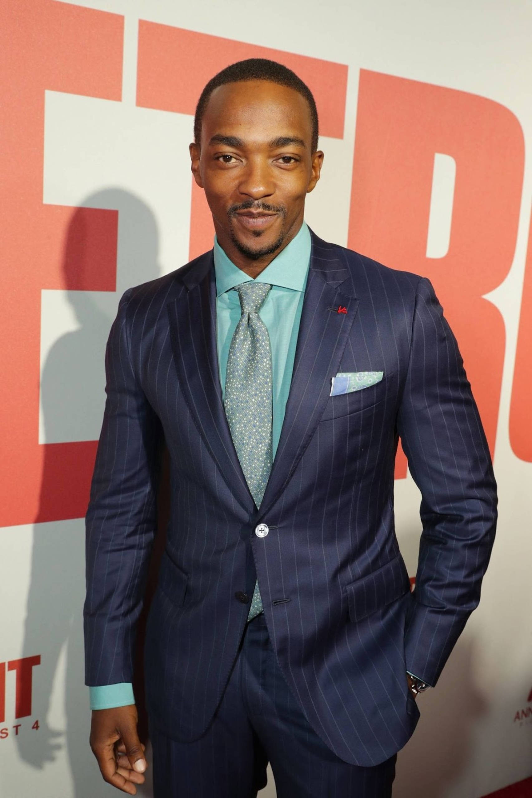 Anthony Mackie 桌布 壁紙 — 一名身穿海軍藍細條紋西裝和青綠色襯衫的男子，在紅白活動背景前自信地擺姿勢。
