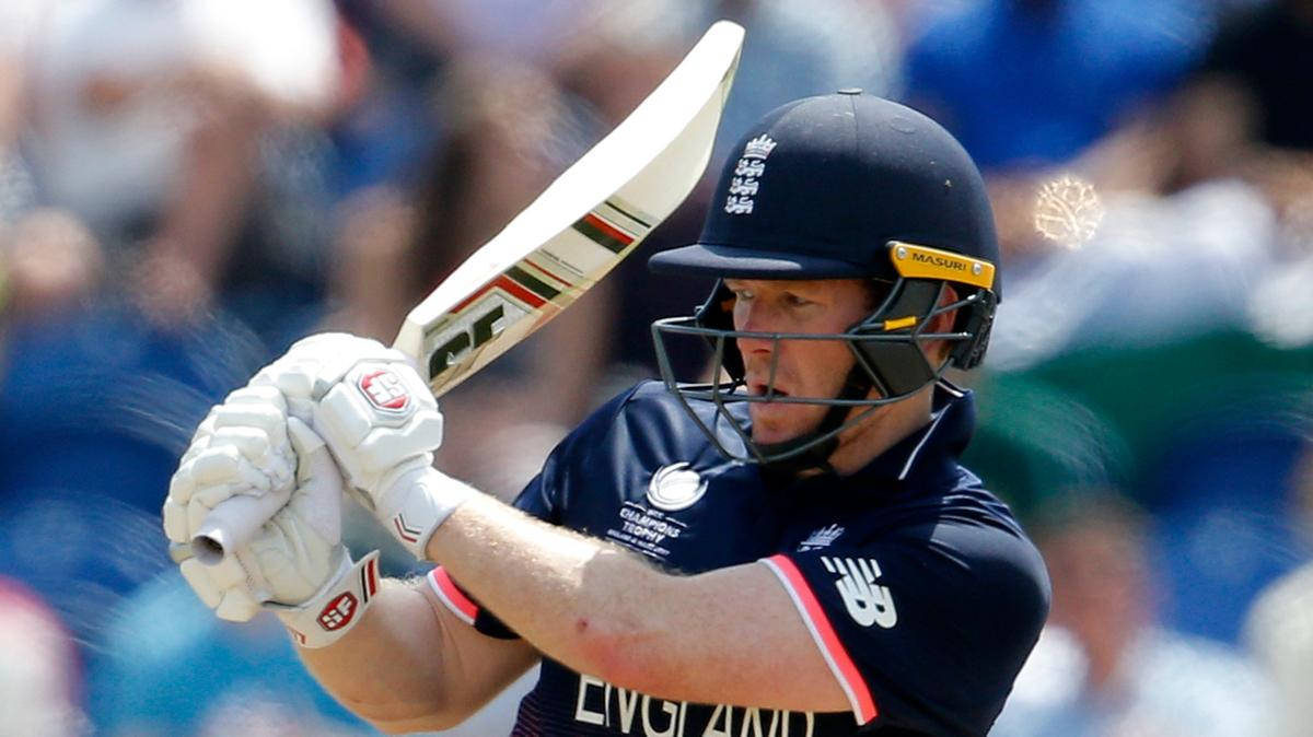 eoin morgan 桌布 壁紙 — 一名身穿海軍藍裝備的英格蘭板球選手在明亮的日間比賽中，專注地揮動球棒。