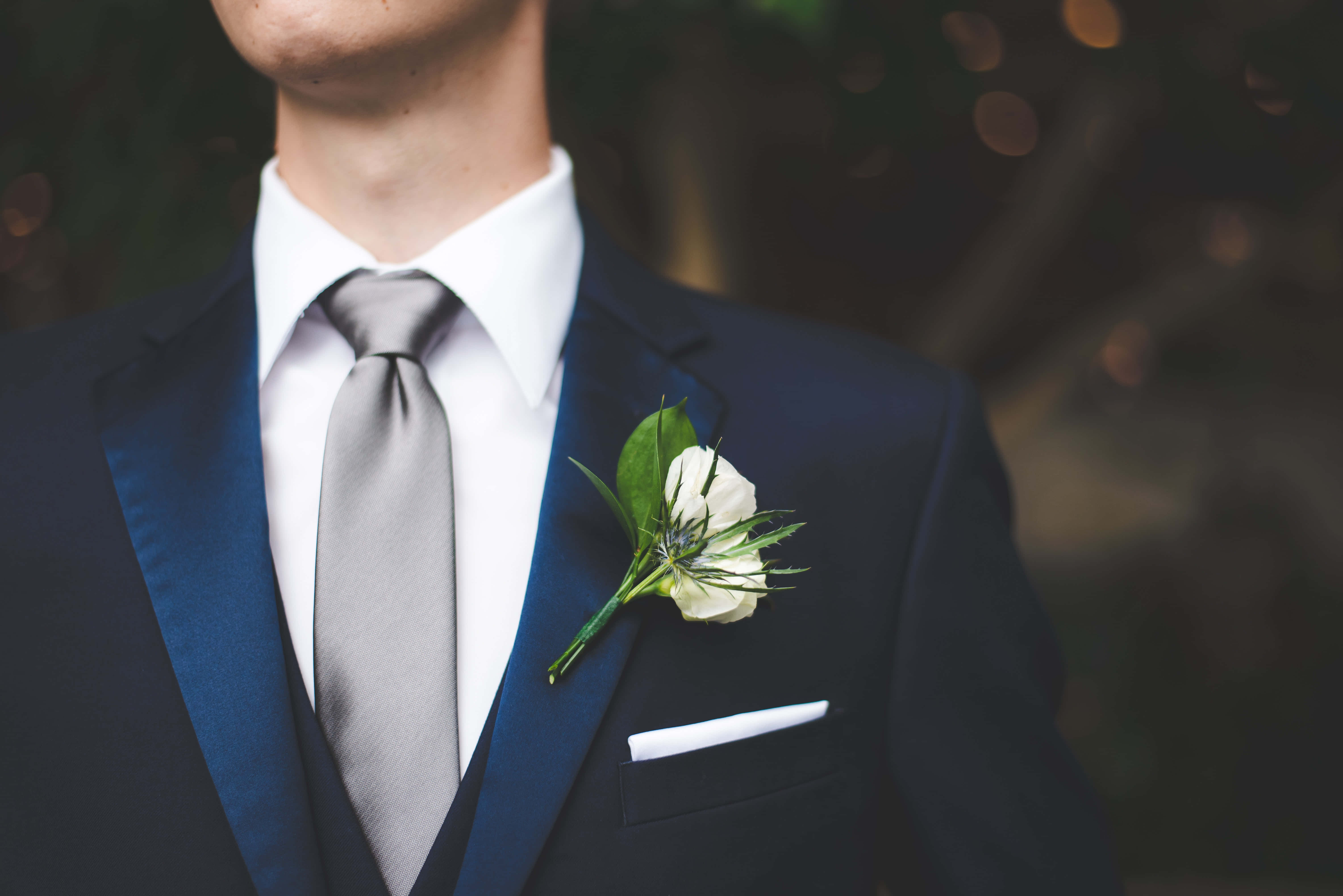 Navy Blue Suit 桌布 壁紙 — 一名身穿海軍藍西裝、銀色領帶和白色襟花的男士特寫，背景是模糊的綠色。