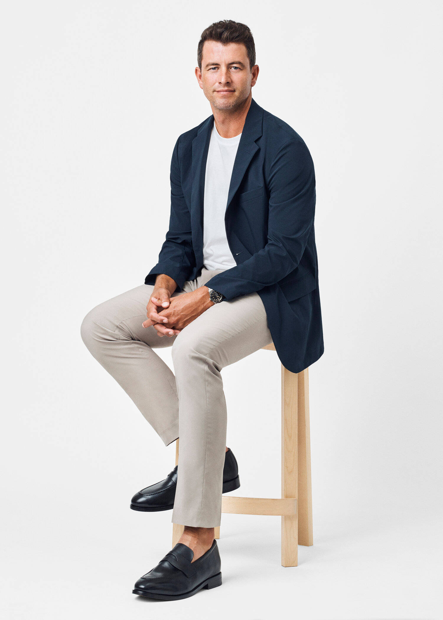 adam scott 桌布 壁紙 — 一名身穿海軍藍西裝外套和米色褲子的男子，隨意地坐在白色背景前的木凳上。