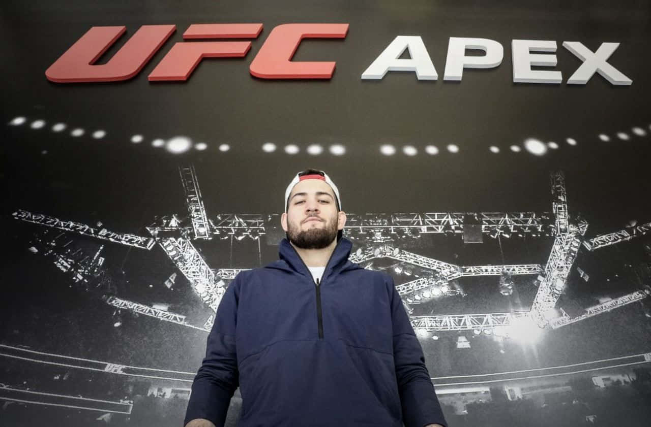 UFC 桌布 壁紙 — 一名身穿海軍藍連帽衫的男子冷靜地站在一個大型UFC APEX標誌前，身後是黑白競技場壁畫。