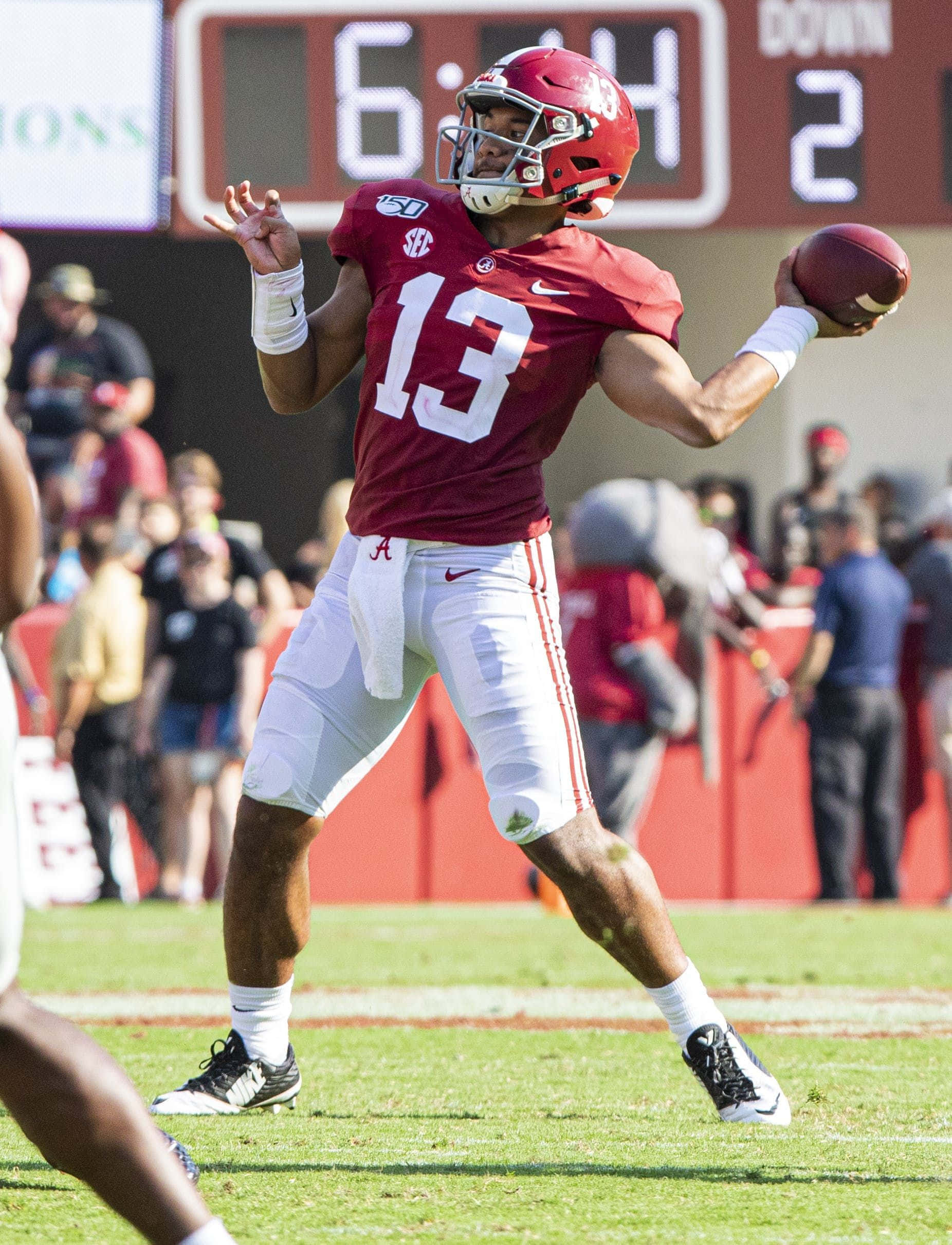 Tua Tagovailoa 桌布 壁紙 — 一名身穿深紅色13號球衣的大學美式足球四分衛在陽光明媚的球場上準備投擲橄欖球，背景有記分牌。