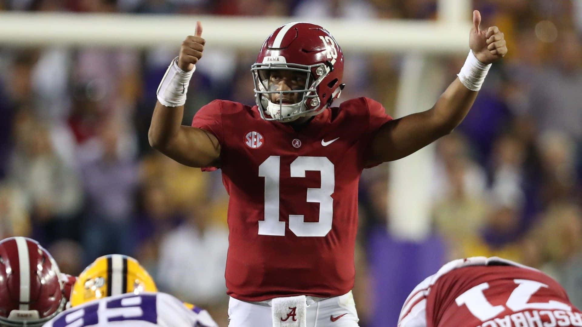 Tua Tagovailoa 桌布 壁紙 — 一名身穿深紅色球衣、號碼13的美式足球員在比賽期間於燈光球場上豎起兩個大拇指。