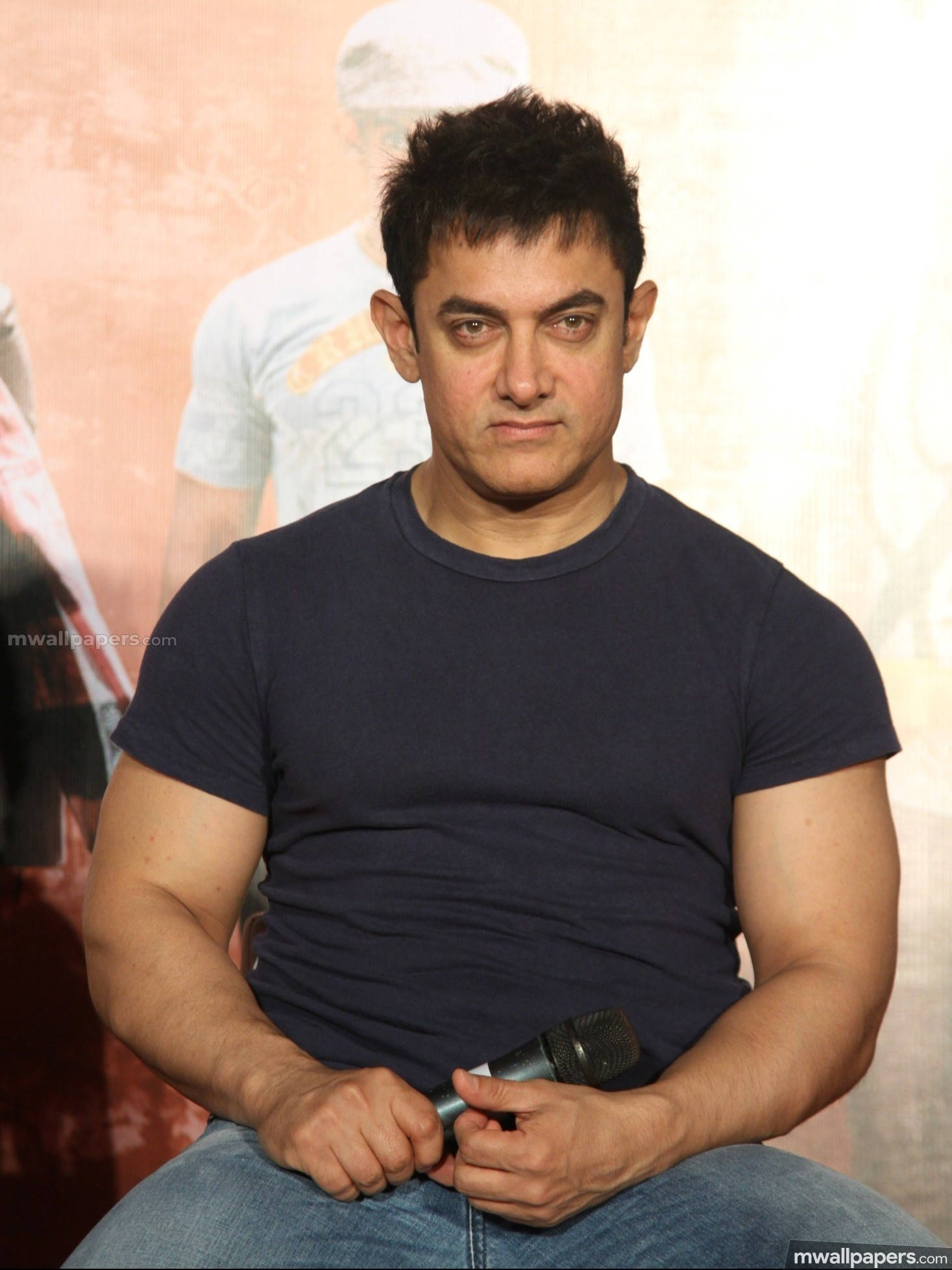 Aamir Khan 桌布 壁紙 — 一名身穿深色T恤和牛仔褲的男子，在模糊的宣傳背景前坐著，手持麥克風。