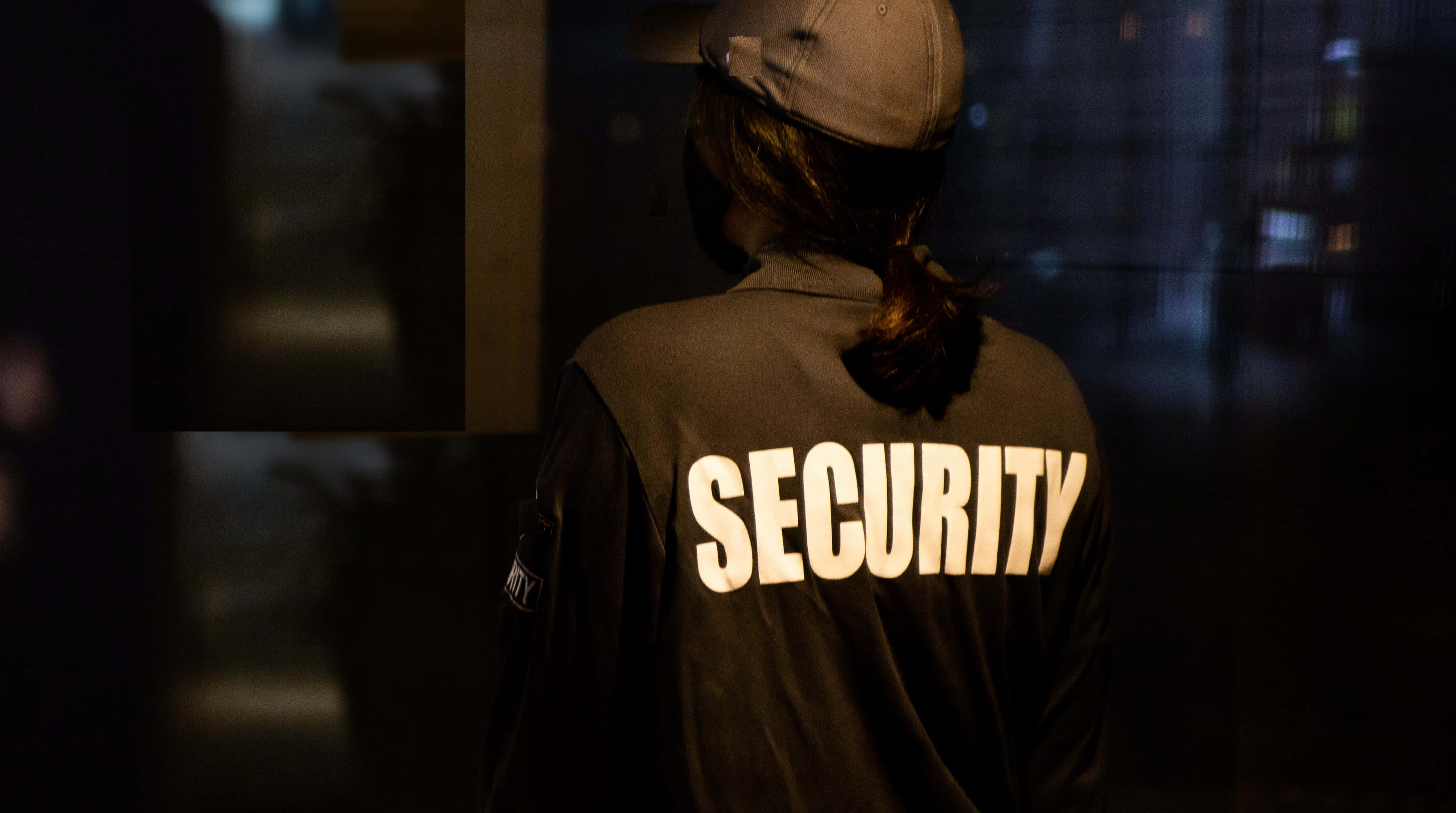 Law Enforcement 桌布 壁紙 — 一名身穿深色制服、帶有發光白色字體的保全人員，夜晚站在光線昏暗的城市環境中。