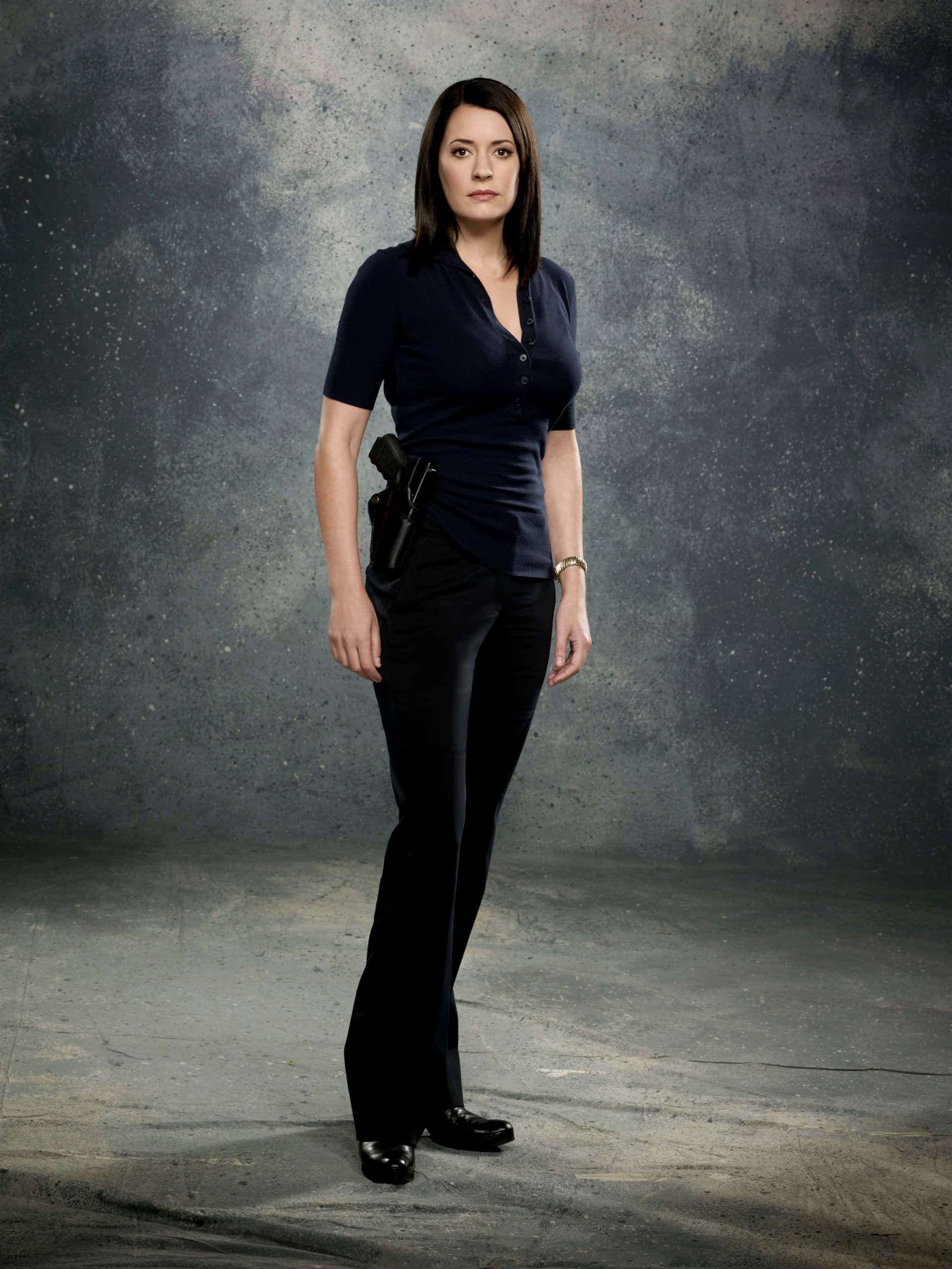 paget brewster 桌布 壁紙 — 一名身穿深色制服的女人站在粗糙紋理的灰色背景前，腰間槍套插著手槍，姿勢穩健。
