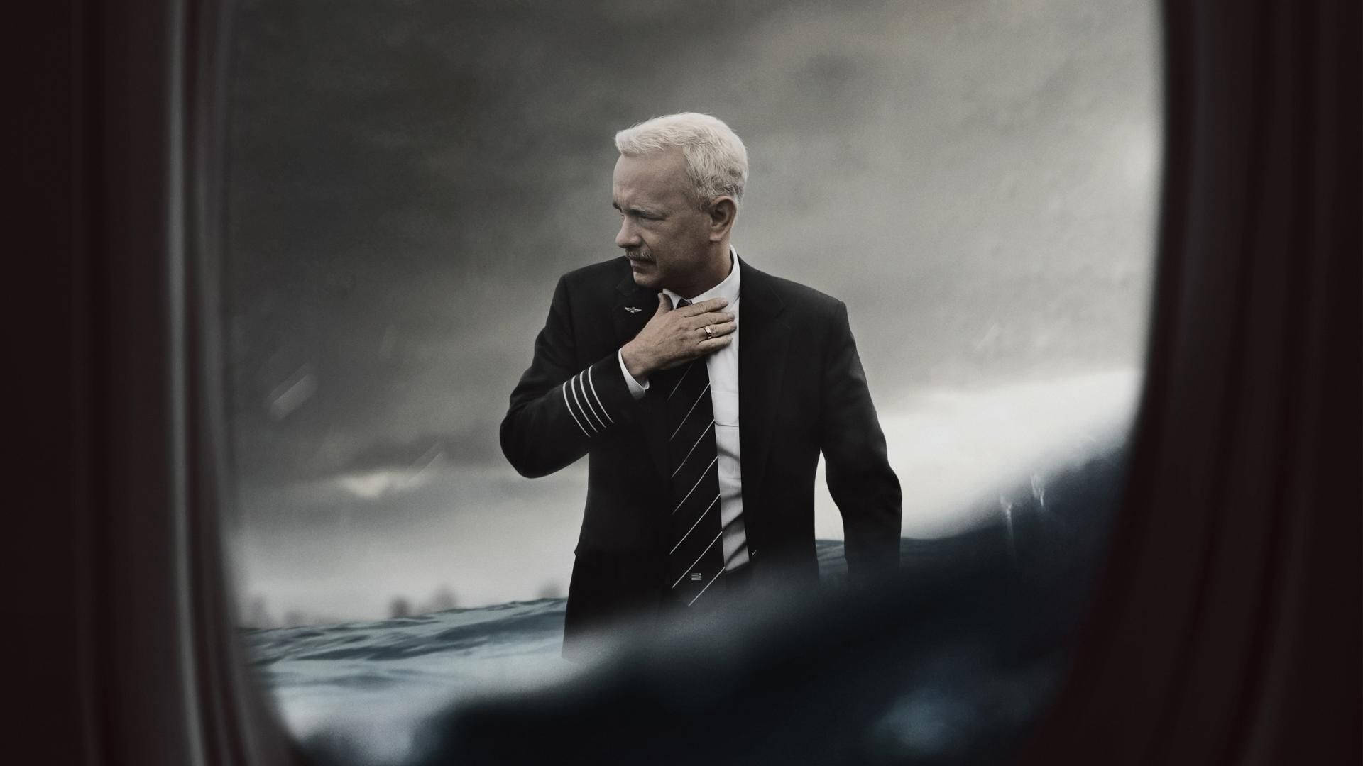 Tom 桌布 壁紙 — 一名身穿深色制服的飛行員在暴風雨的海邊莊嚴地站著，透過窗戶和模糊的前景窗簾觀看。
