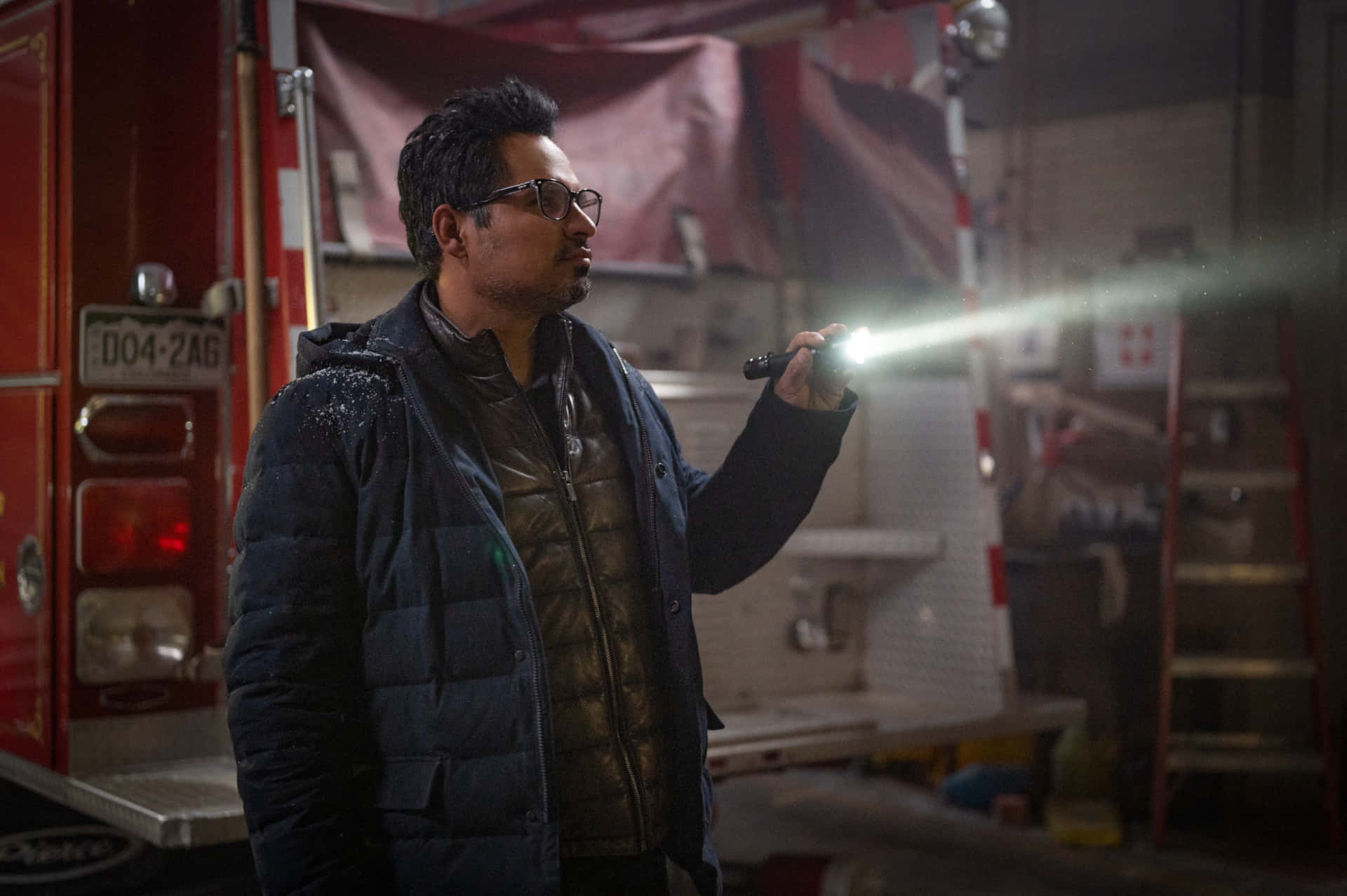 Michael Peña 桌布 壁紙 — 一名身穿深色外套、戴眼鏡的男子在光線昏暗的消防局內用手電筒照明，附近有一輛紅色消防車。