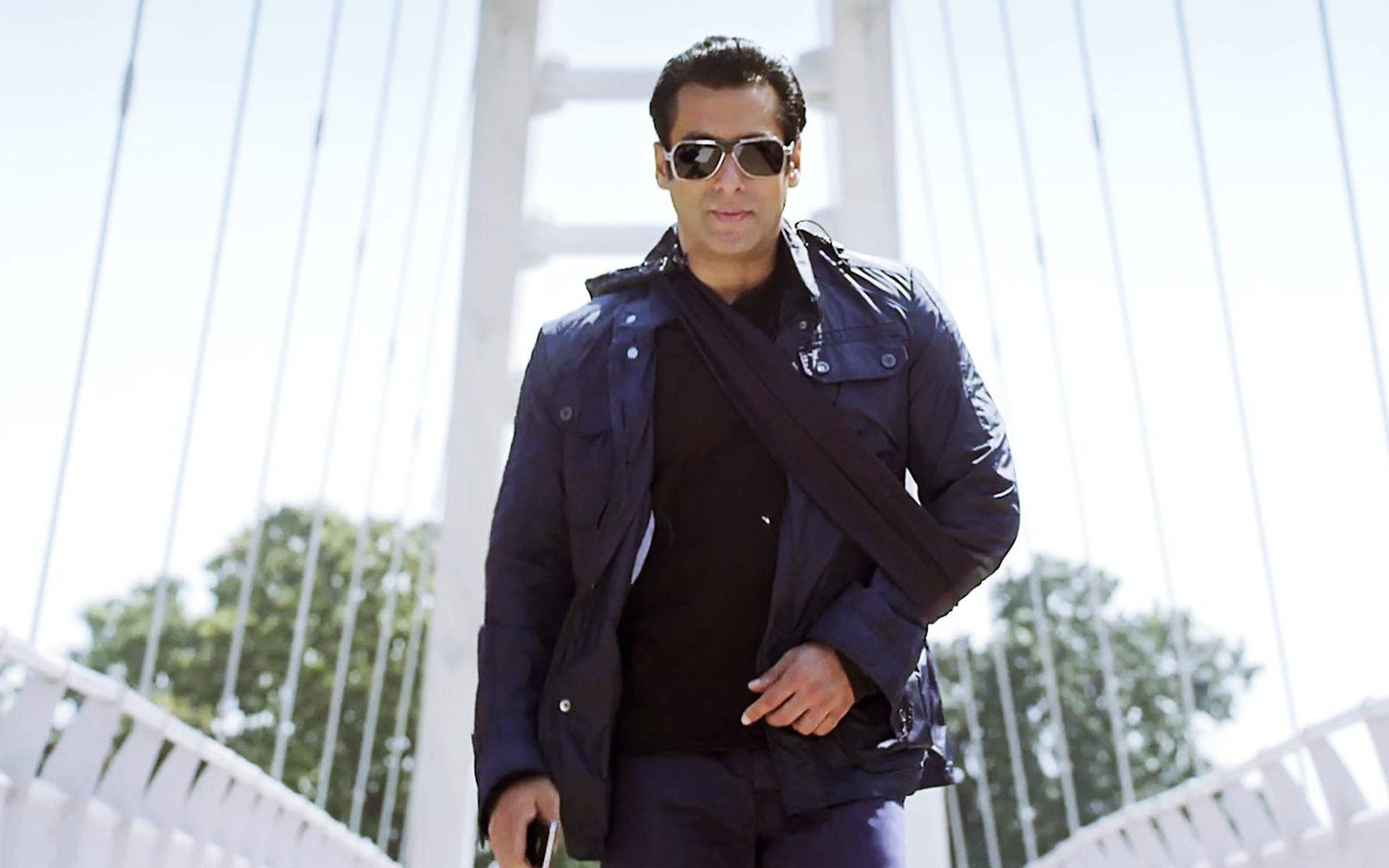salman khan HD 桌布 壁紙 — 一名身穿深色外套和太陽眼鏡的男子，在明亮的日光下自信地走過一座現代白色吊橋。
