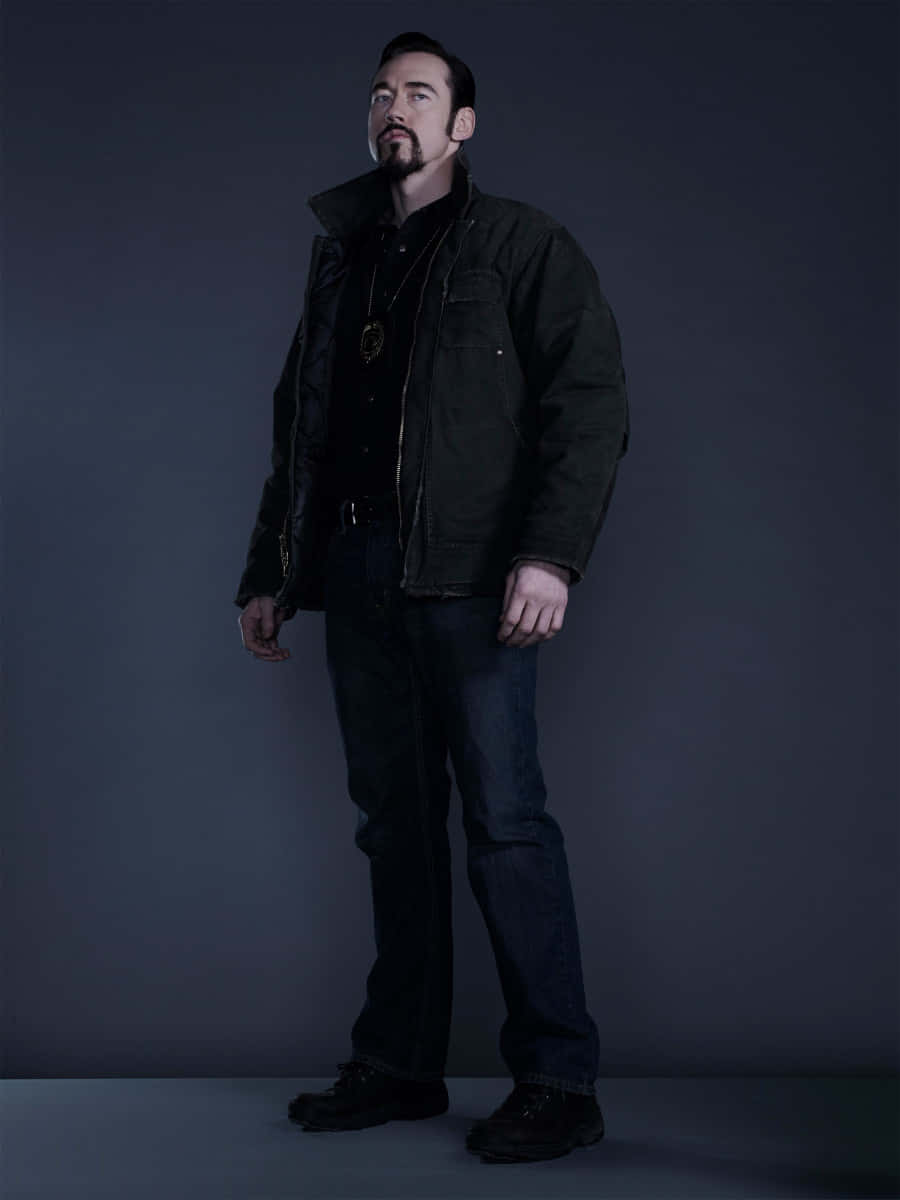 Kevin Durand 桌布 壁紙 — 一名身穿深色外套和牛仔褲的男子站在帶有戲劇性燈光的陰鬱灰色工作室背景前。