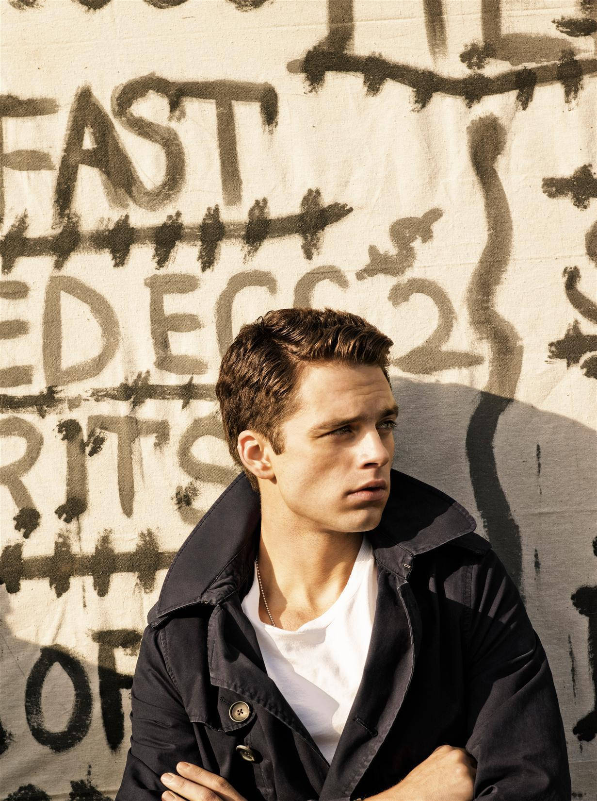 Sebastian 桌布 壁紙 — 一名身穿深色外套的年輕男子站在陽光照射的塗鴉牆前，牆上有粗體黑色標籤。