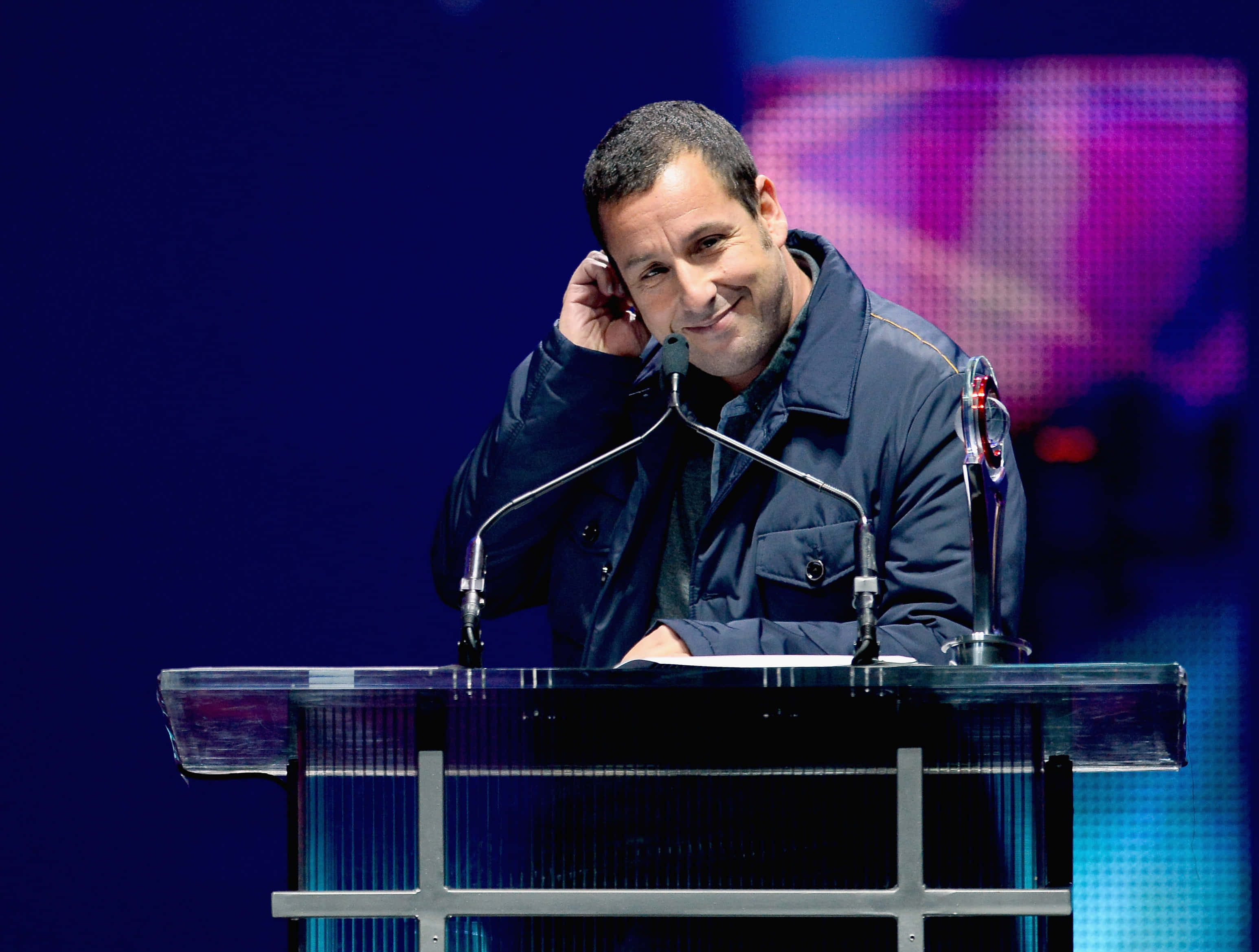 Adam Sandler 桌布 壁紙 — 一名身穿深色外套的男子在充滿活力的藍色和紫色舞台燈光下，靠在透明講台上溫暖地微笑。