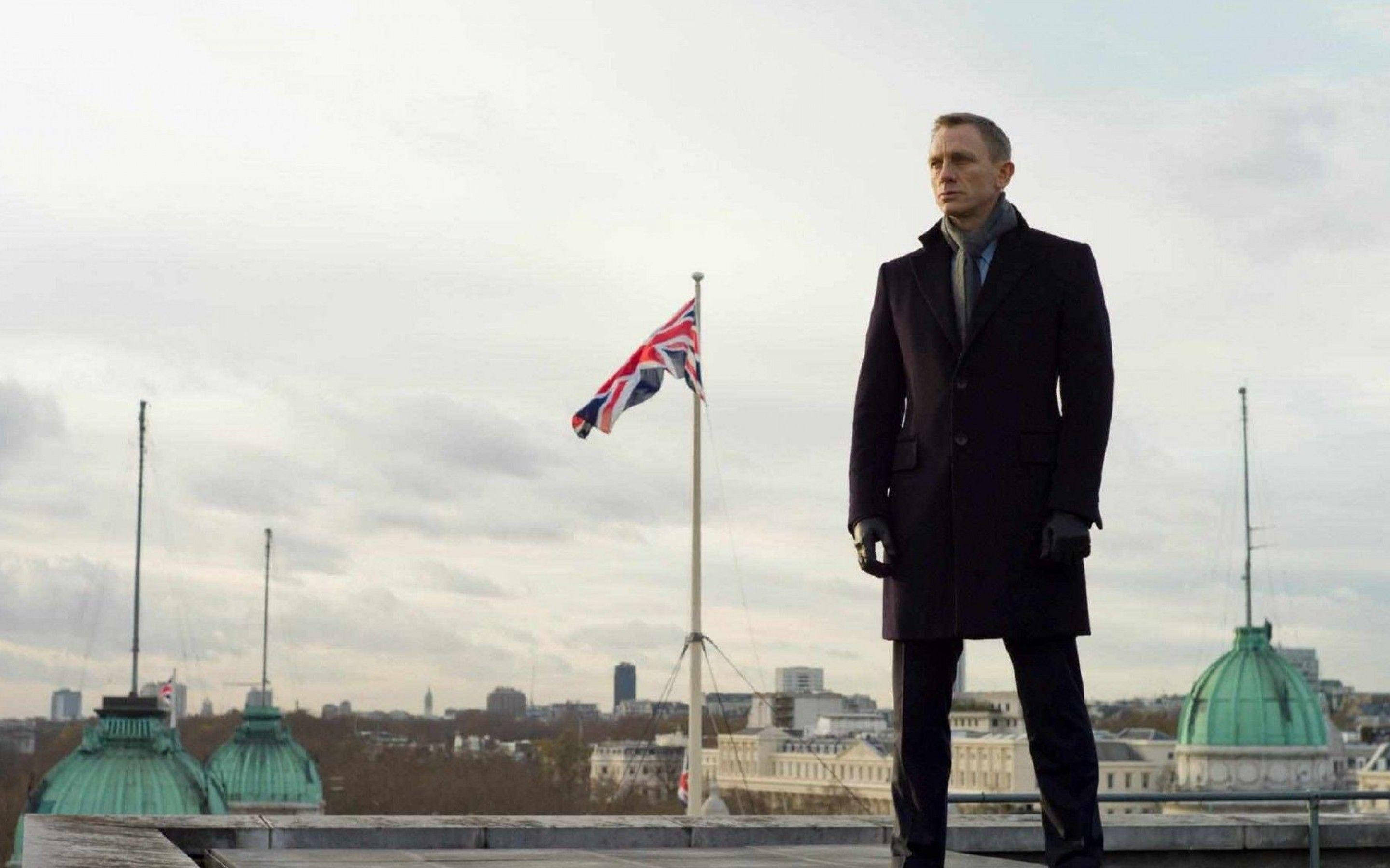 Bond 桌布 壁紙 — 一名身穿深色外套的男子莊嚴地站在屋頂上，英國國旗在陰雲密布的倫敦天際線中飄揚。