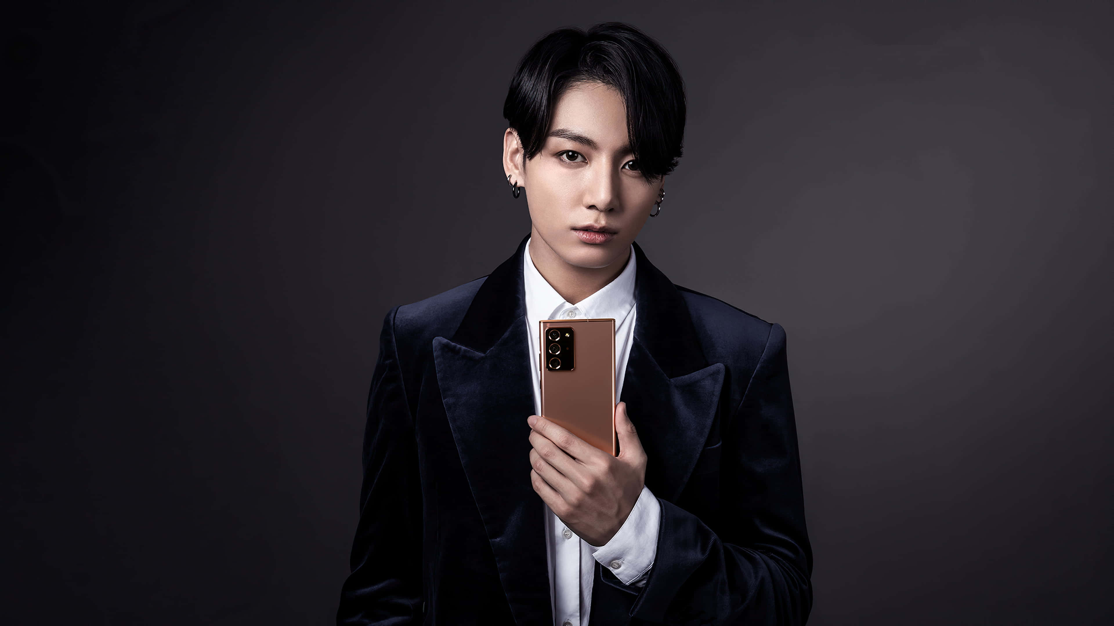 Jungkook 桌布 壁紙 — 一名身穿深色天鵝絨西裝的男子，在陰鬱的灰色背景下手持玫瑰金智慧型手機，目光直接而強烈。