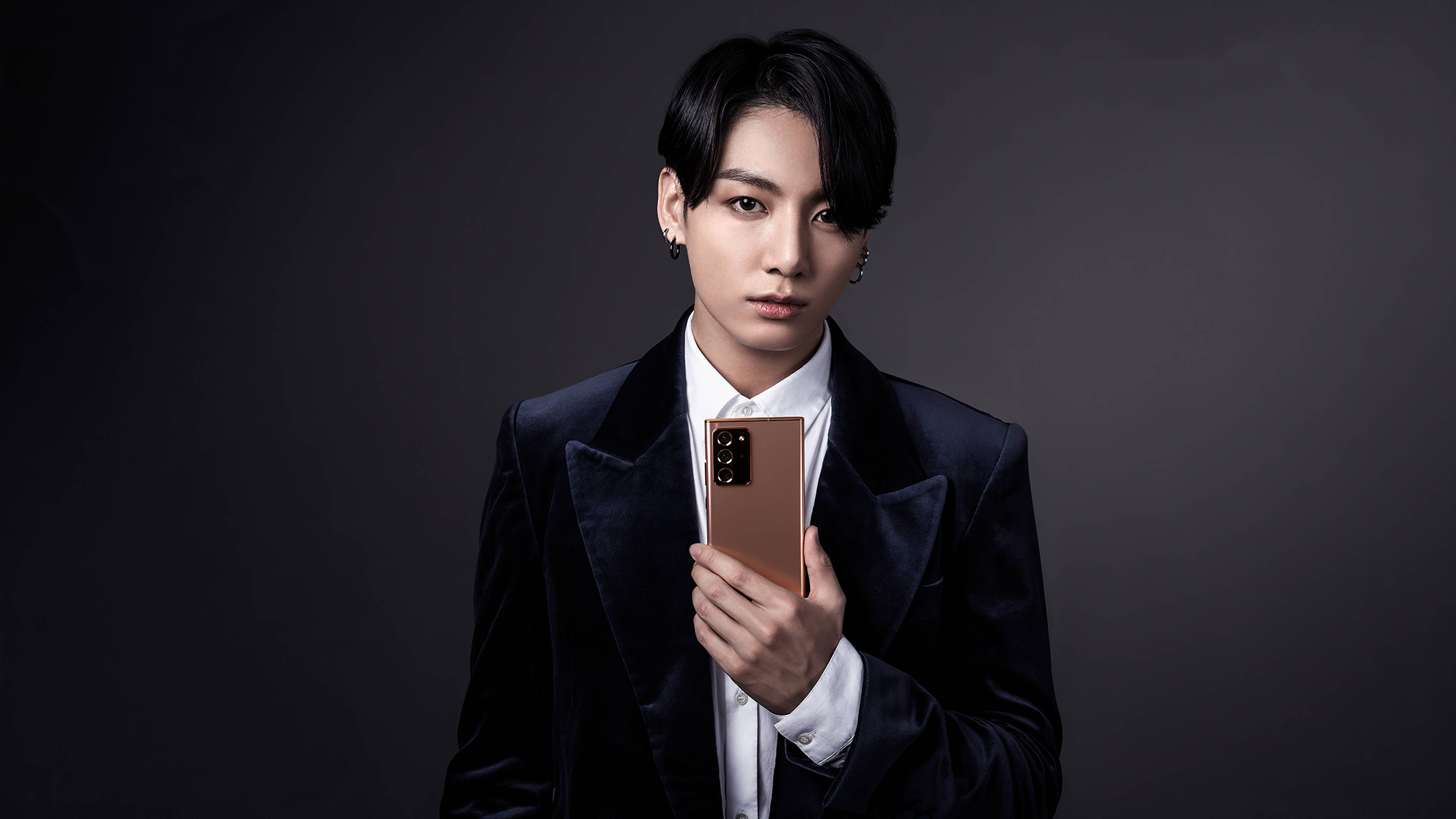 美學 BTS 桌布 壁紙 — 一名身穿深色天鵝絨西裝的男子，在柔和的灰色背景前手持玫瑰金手機，眼神直接而強烈。