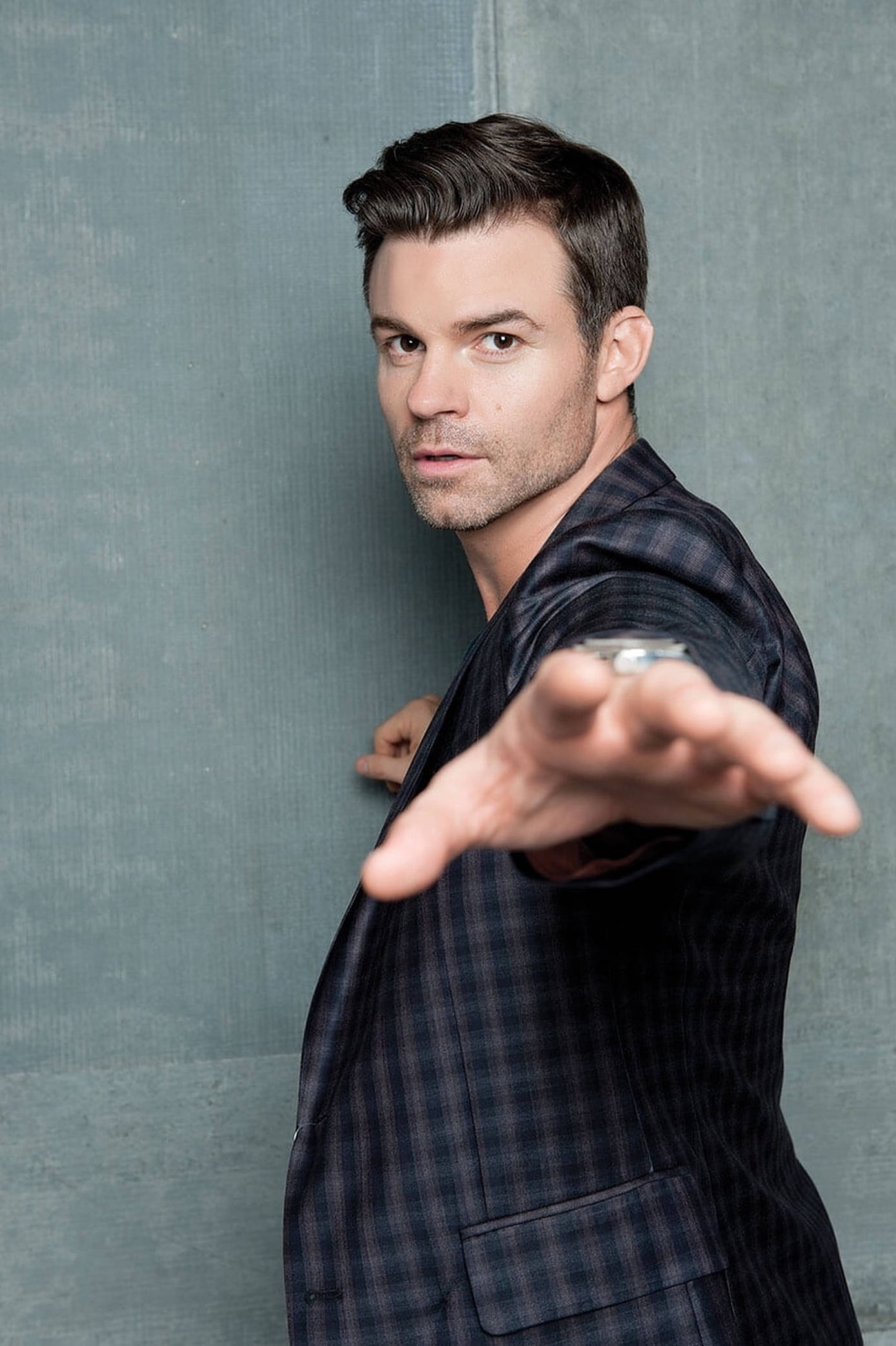 Daniel Gillies 桌布 壁紙 — 一名身穿深色格紋西裝外套的男子，對著有紋理的灰色牆壁伸出手，目光直接而強烈。