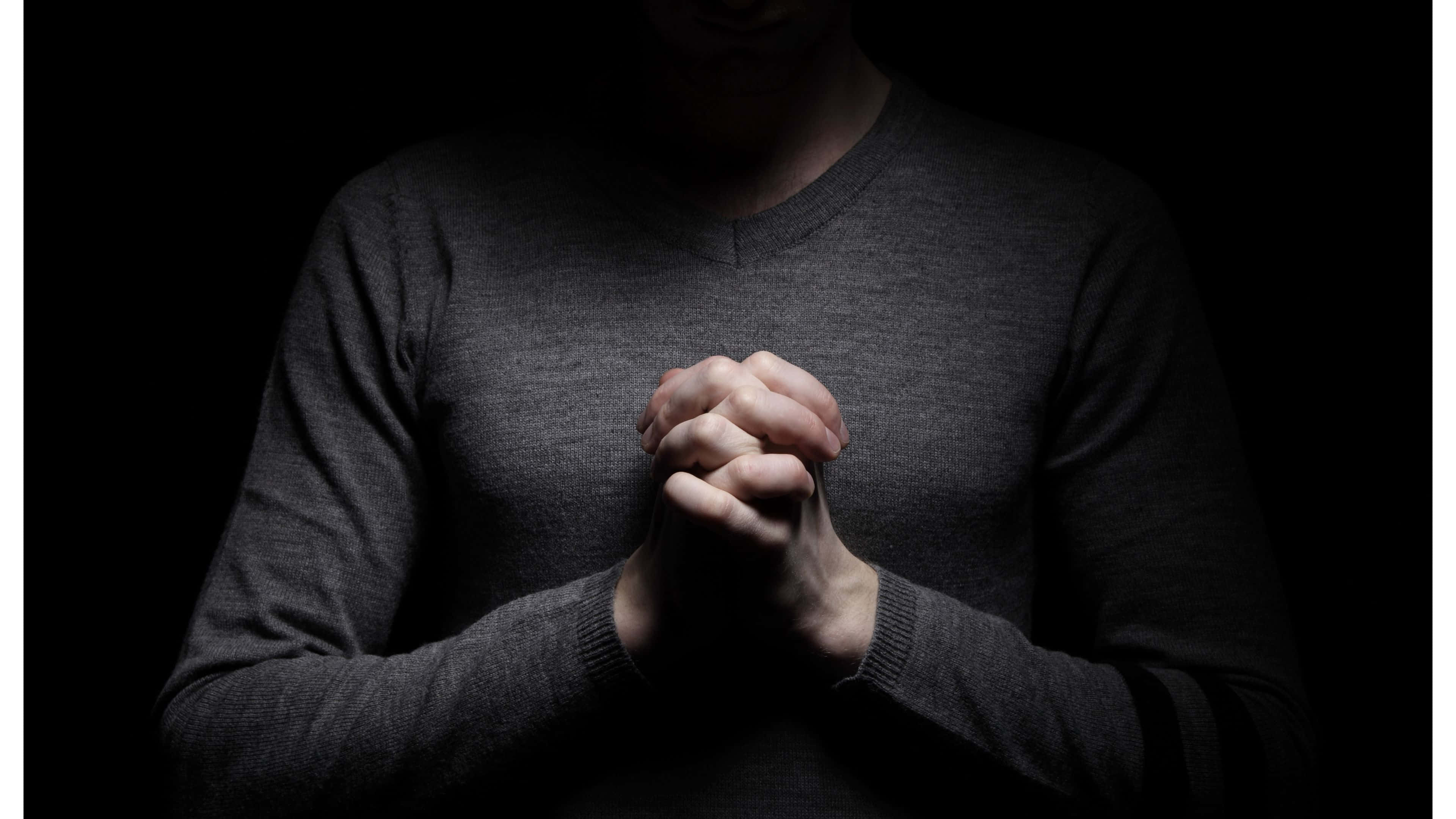 Praying 桌布 壁紙 — 一名身穿深色毛衣的人，在戲劇性聚光燈下，於黑色背景前合十祈禱。