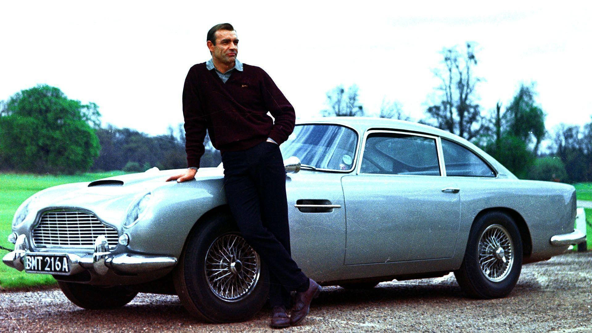 Bond 桌布 壁紙 — 一名身穿深色毛衣的男子自信地靠在一輛銀色Aston Martin DB5上，背景是碎石路和綠樹。