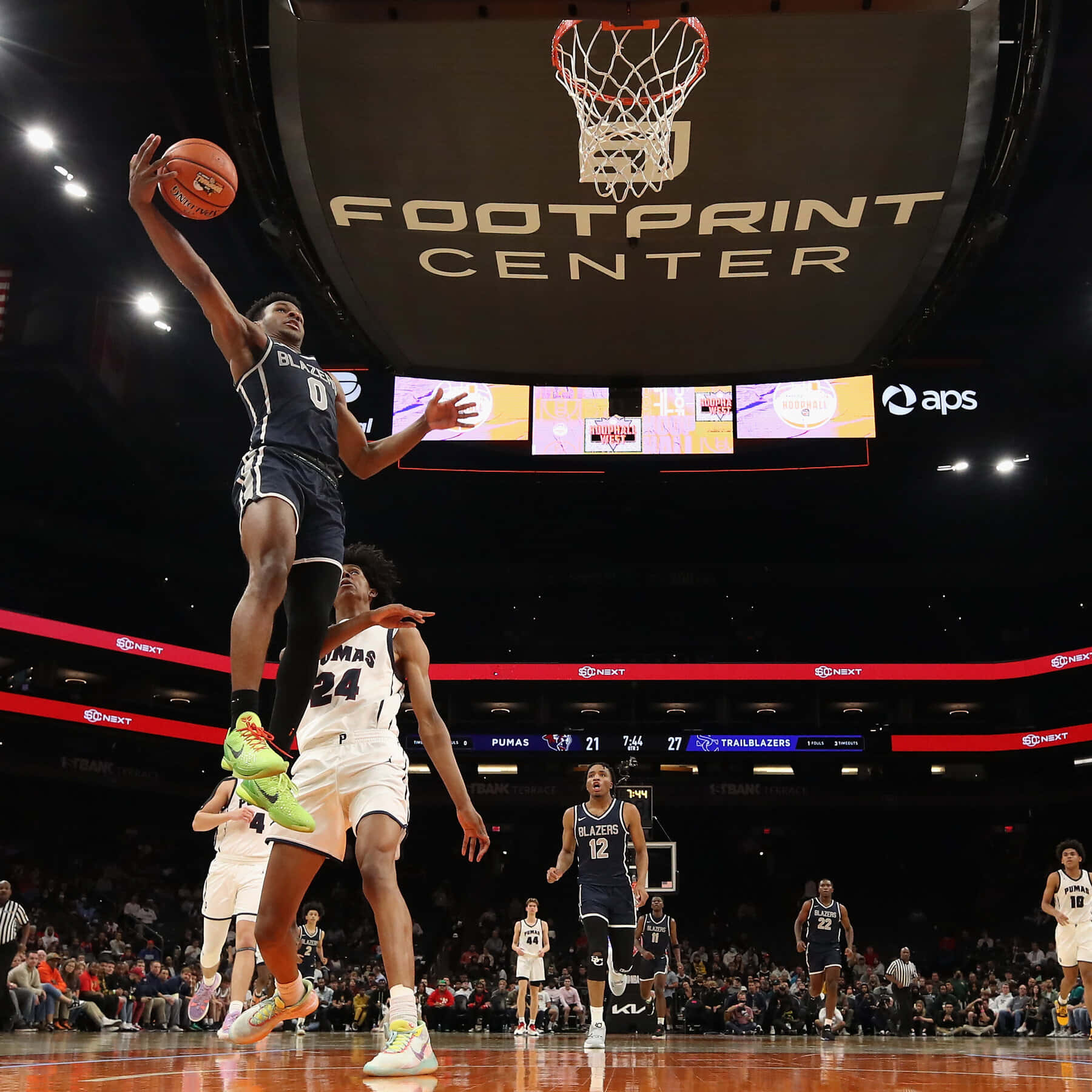Bronny James 桌布 壁紙 — 一名身穿深色球衣的籃球運動員高高躍起準備灌籃，而一名白色球衣的防守球員在場館燈光下於下方觀看。