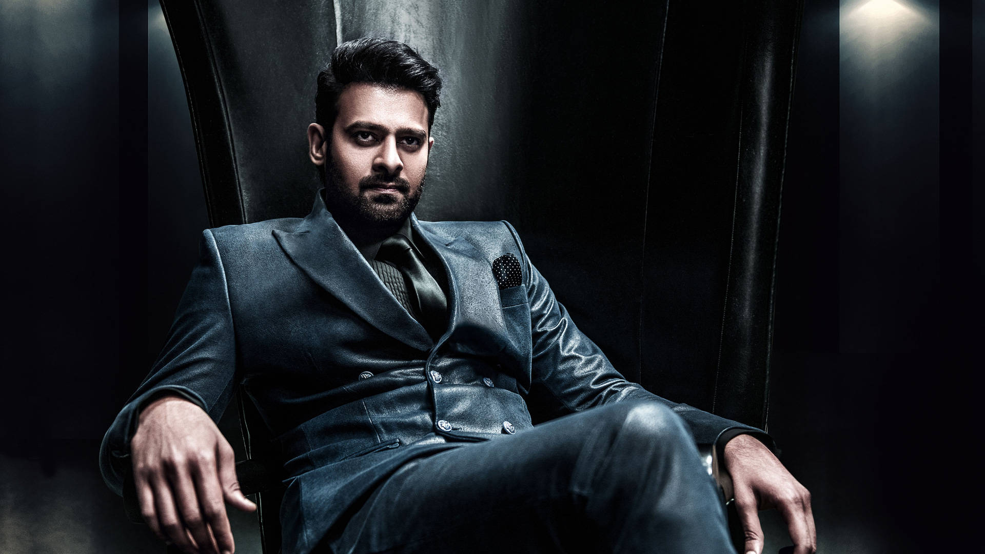 Prabhas HD 桌布 壁紙 — 一名身穿深色皮套裝的男子在戲劇性的聚光燈下自信地坐在類似王座的椅子上。