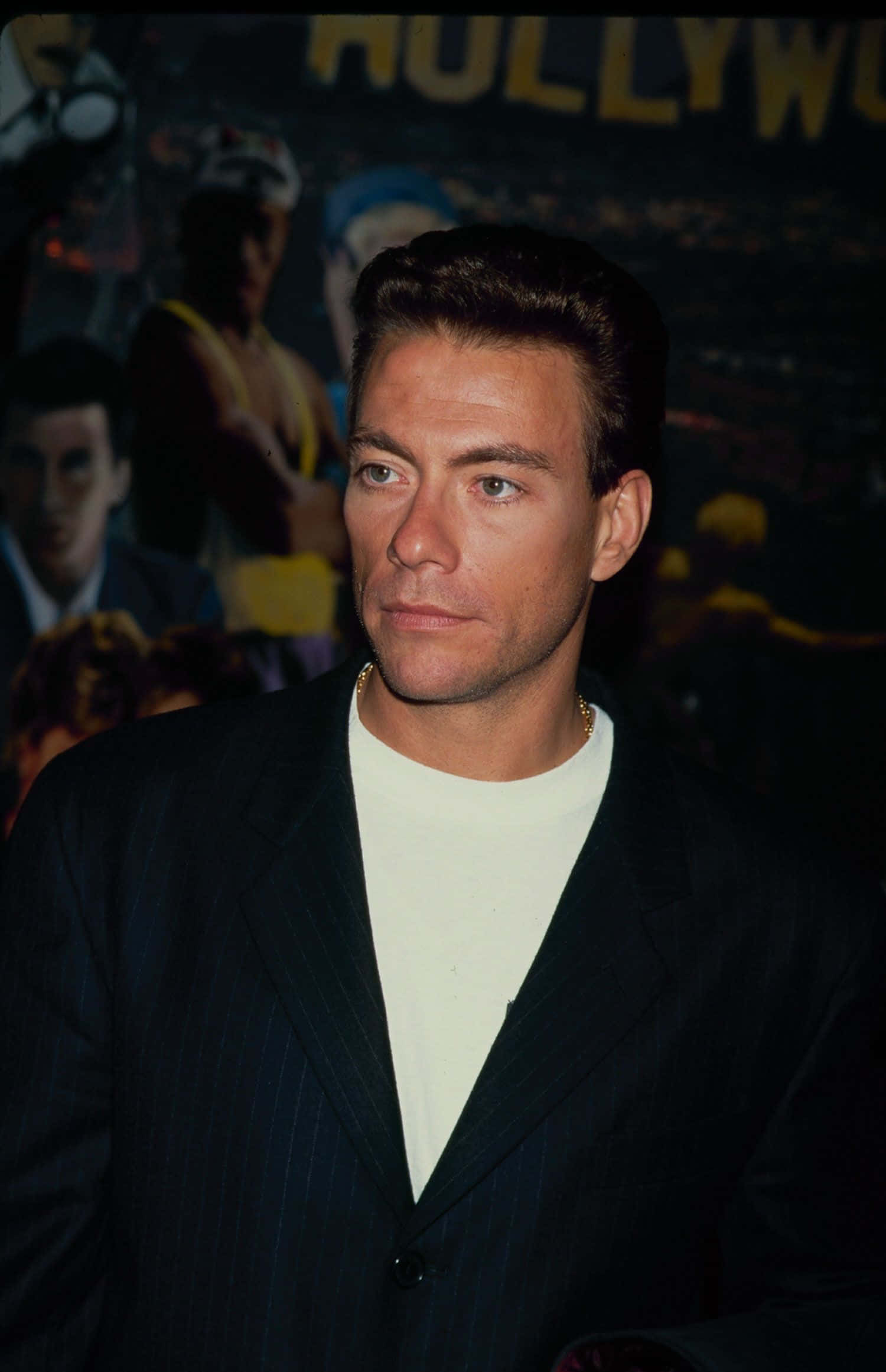 jean claude van damme 桌布 壁紙 — 一名身穿深色細條紋西裝和白色T恤的男子，站在模糊的好萊塢背景前，周圍有溫暖的環境燈光。