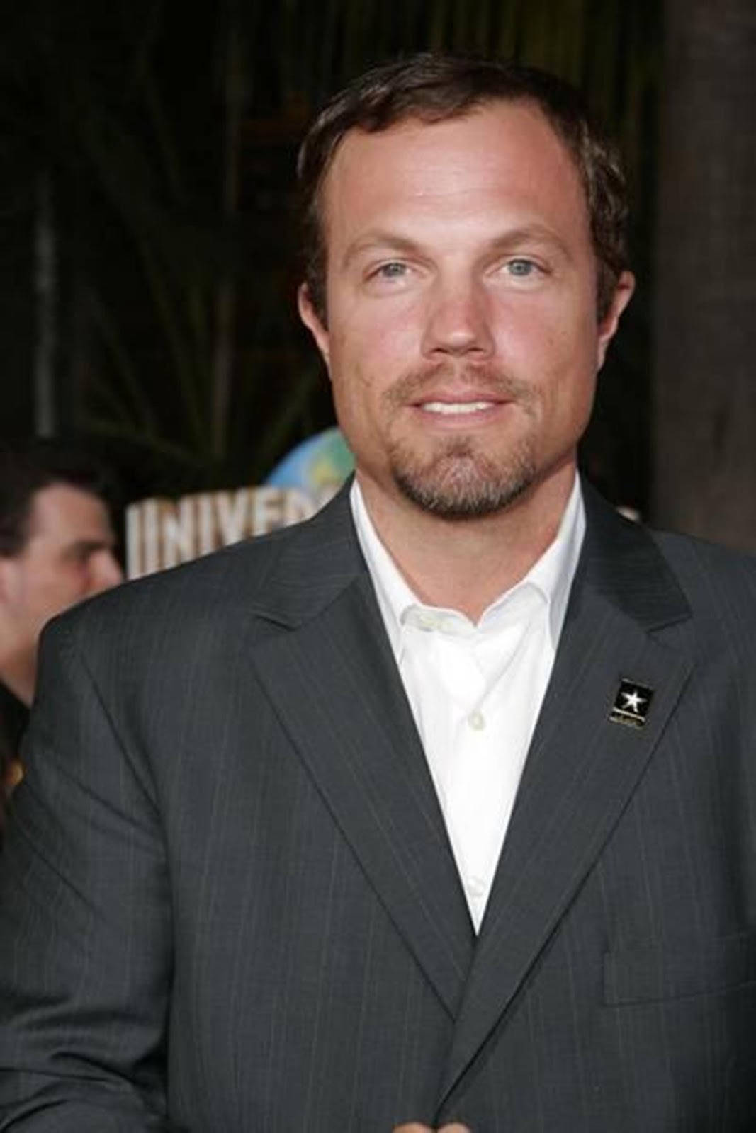 adam baldwin 桌布 壁紙 — 一名身穿深色細條紋西裝和白色襯衫的男子在環球影業活動中對著鏡頭微笑，身後有模糊的樹葉和標牌。