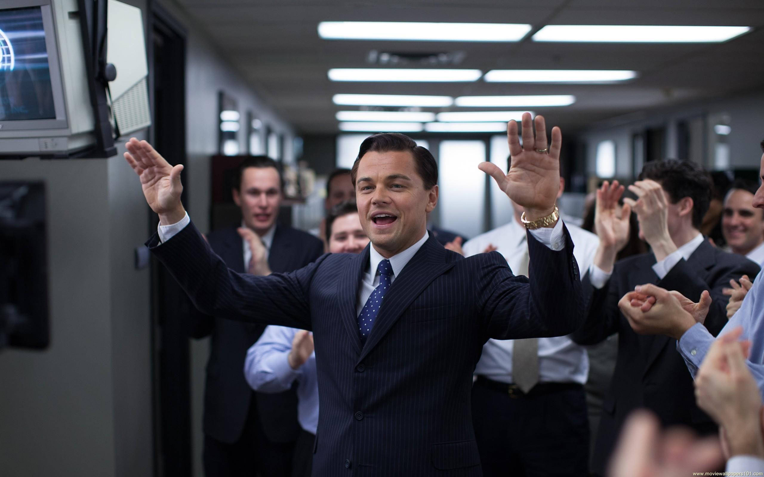 Wolf Of Wall Street 桌布 壁紙 — 一名身穿深色細條紋西裝的男子在光線明亮的辦公室走廊中高舉雙手，慶祝勝利，周圍同事歡呼。