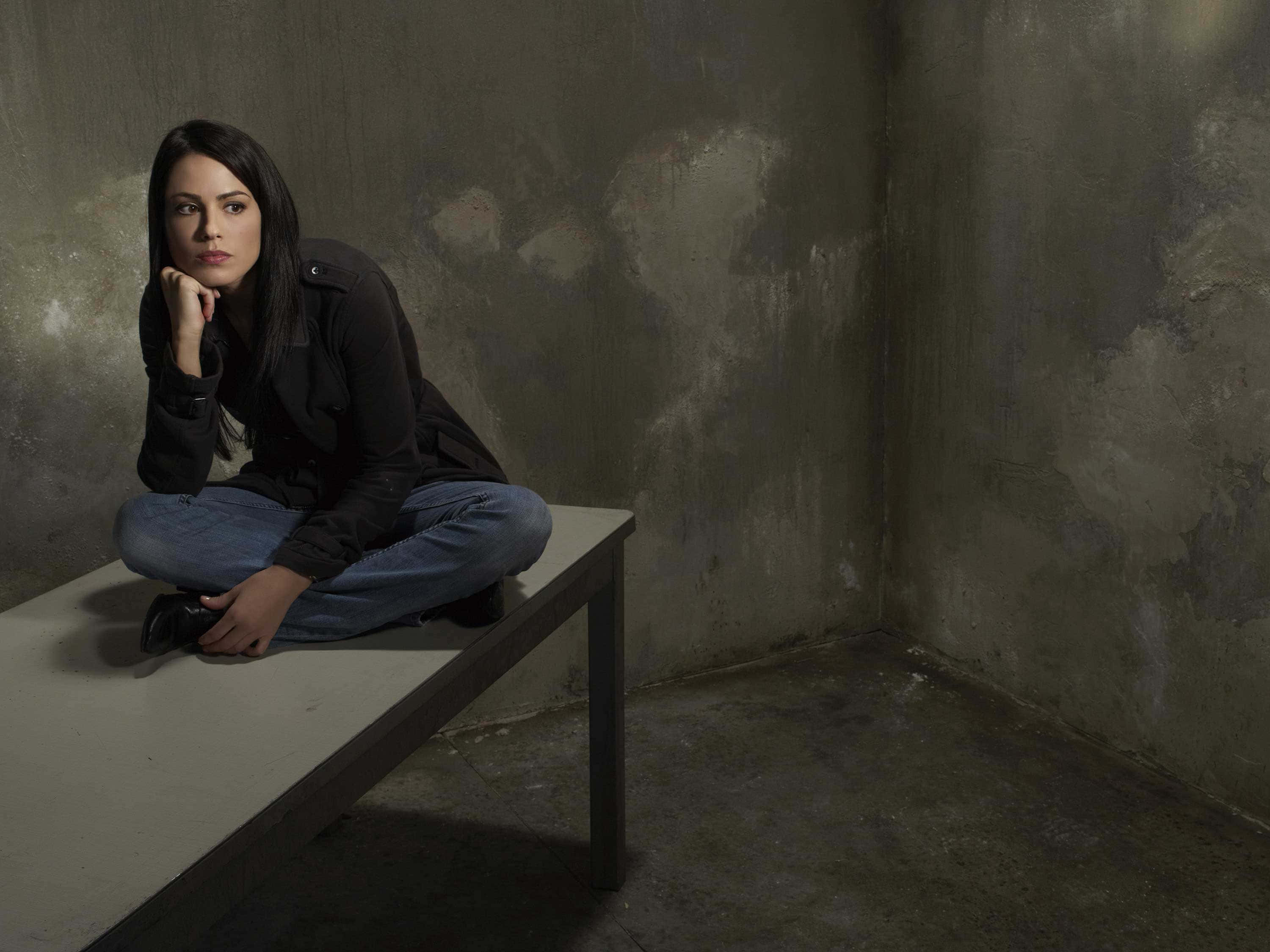 Michelle Borth 桌布 壁紙 — 一名身穿深色衣服的女子在光線昏暗的房間裡，靠著風化的混凝土牆，若有所思地坐在長椅上。