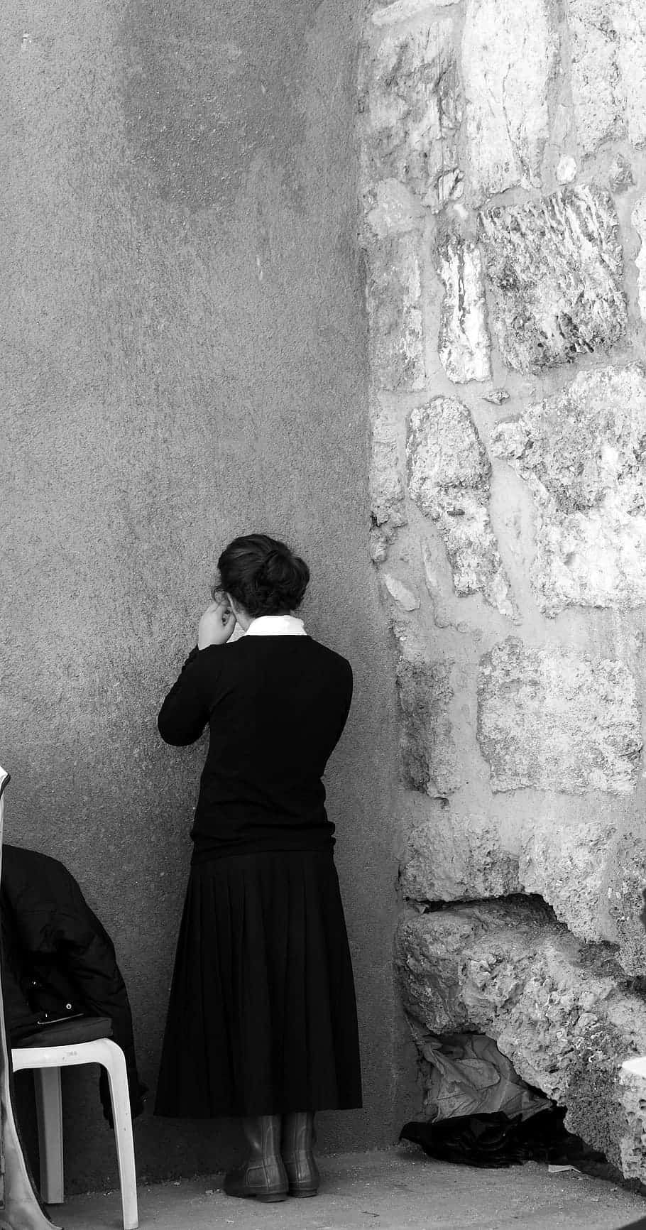 Praying 桌布 壁紙 — 一名身穿深色衣服的女子背對鏡頭，靠著粗糙的石牆，呈現鮮明的黑白構圖。