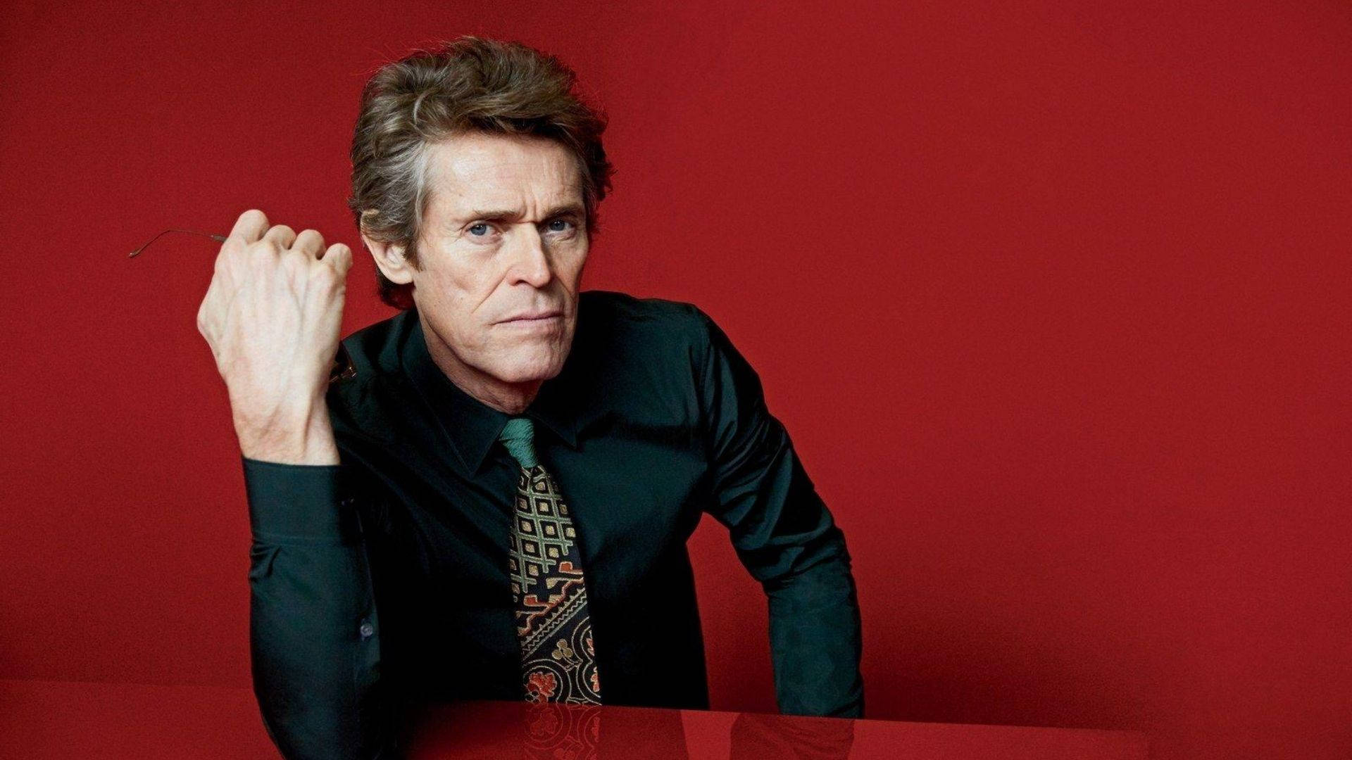 Willem Dafoe 桌布 壁紙 — 一名身穿深色襯衫和圖案領帶的男子，在鮮紅色背景前將一個薄物體靠近耳朵。