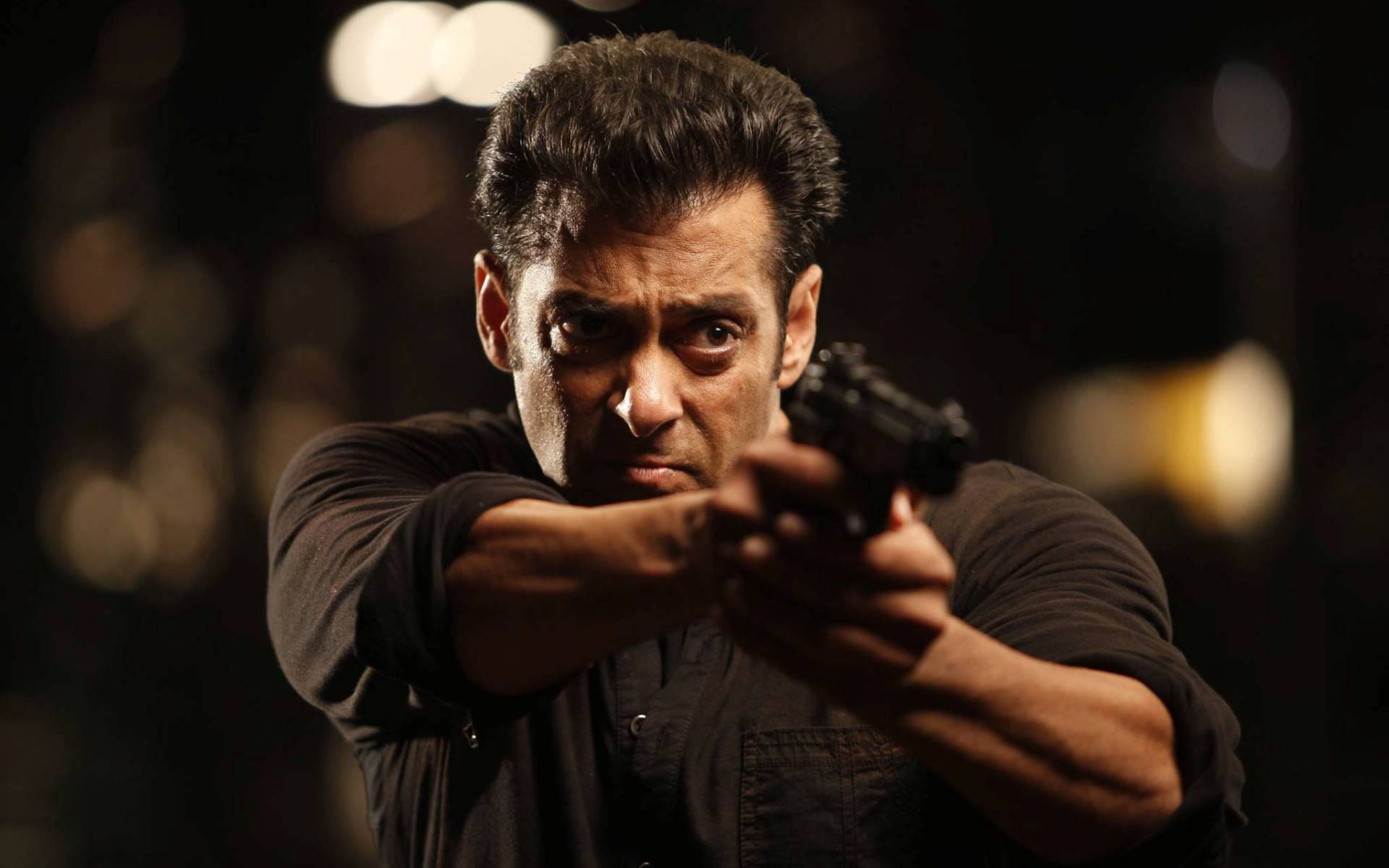 Salman Khan 桌布 壁紙 — 一名身穿深色襯衫的男子，在模糊的夜間背景和散景燈光下，專注地瞄準手槍。