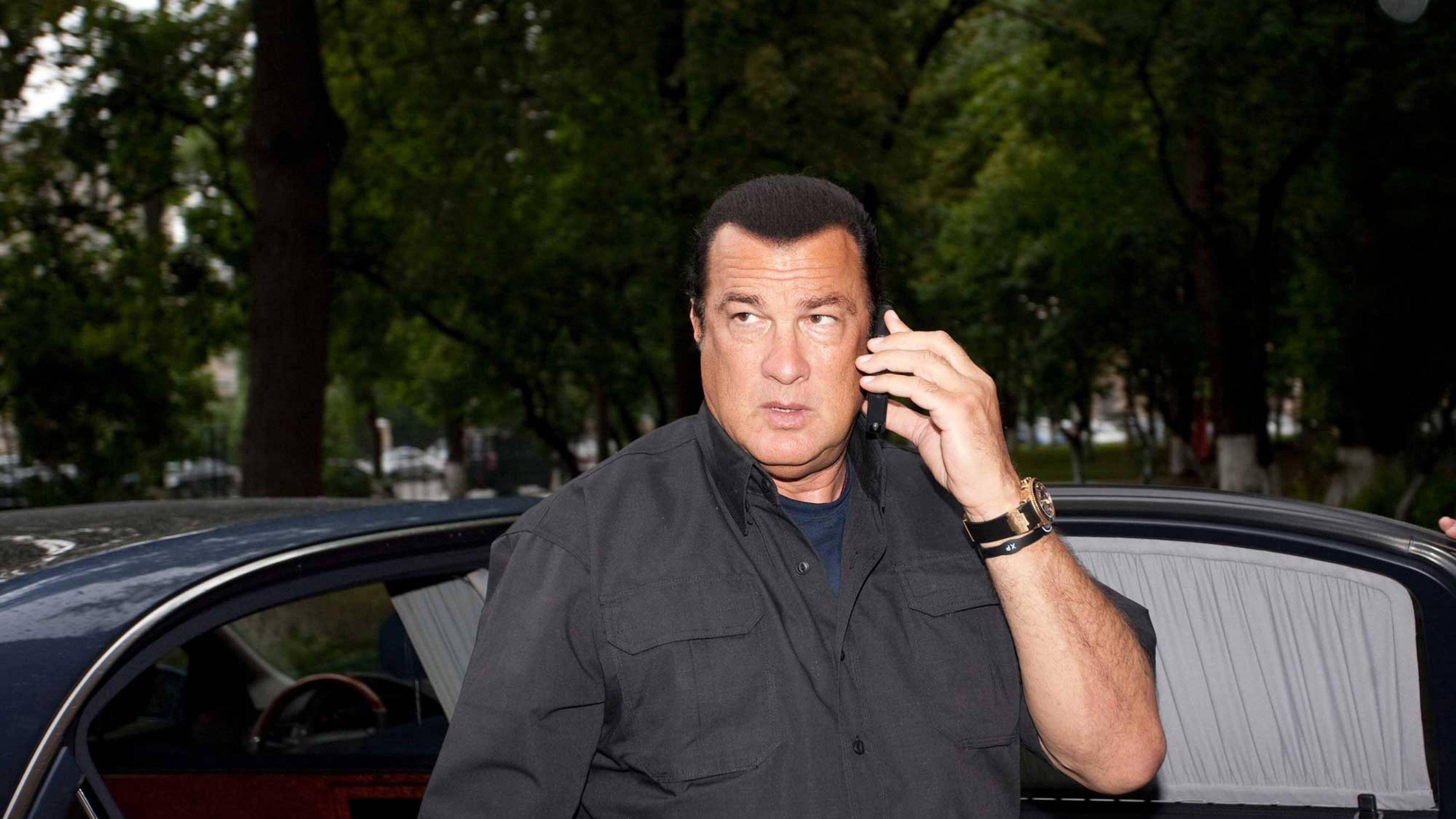 Steven Seagal 桌布 壁紙 — 一名身穿深色襯衫的男子在黑色汽車旁講電話，背景有樹木。