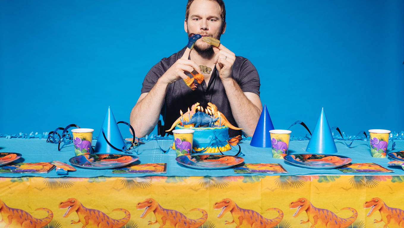 Chris Pratt 桌布 壁紙 — 一名身穿深色襯衫的男子坐在色彩繽紛的恐龍主題生日派對桌前，桌上放有蛋糕和派對帽，背景為純藍色。