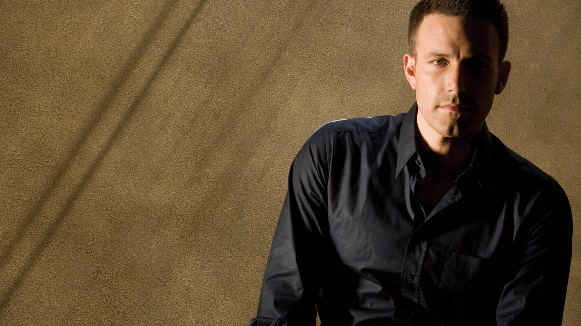 Ben Affleck 桌布 壁紙 — 一名身穿深色襯衫的男子靠著帶有紋理的牆壁坐下，牆面上有對角線陰影投射。