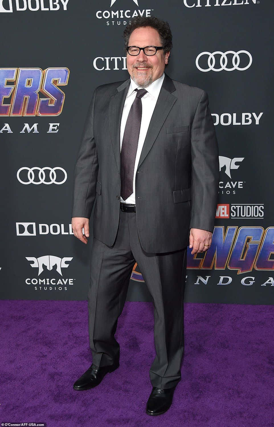 Jon Favreau 桌布 壁紙 — 一名身穿深色西裝、戴眼鏡的男子在Marvel Studios活動的紫色地毯上微笑，身後有贊助商標誌。