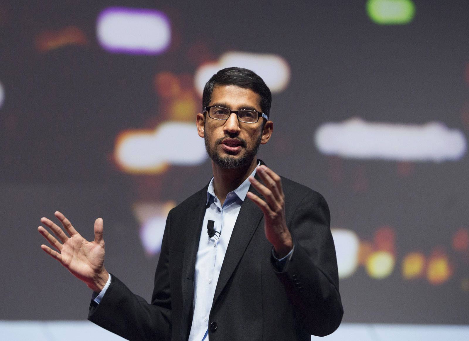 google 桌布 壁紙 — 一名身穿深色西裝、戴眼鏡的男子在科技會議舞台上說話時做手勢，身後有模糊的彩色燈光。